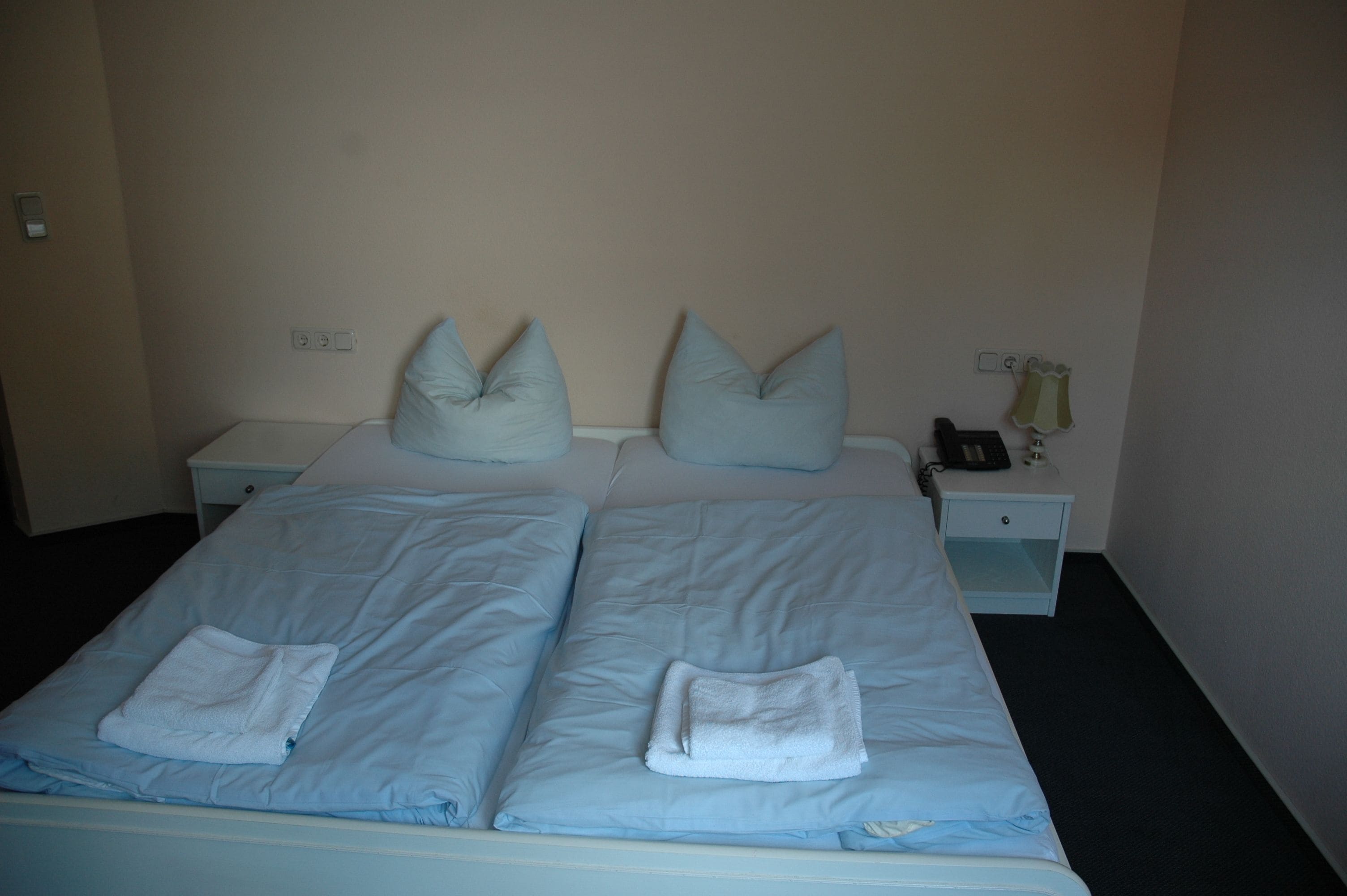 A twin bed with two pillows and a folded towel. There is a nightstand with a lamp and a phone.