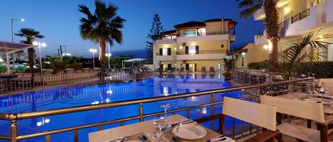 Un área de comedor junto a la piscina con mesas preparadas para dos, con vistas a una piscina y un edificio de varios pisos al fondo.