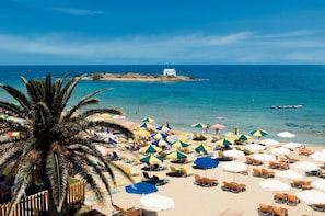 Een strand met vele ligbedden en parasols, een palmboom en een klein eiland met een wit gebouw op de achtergrond.