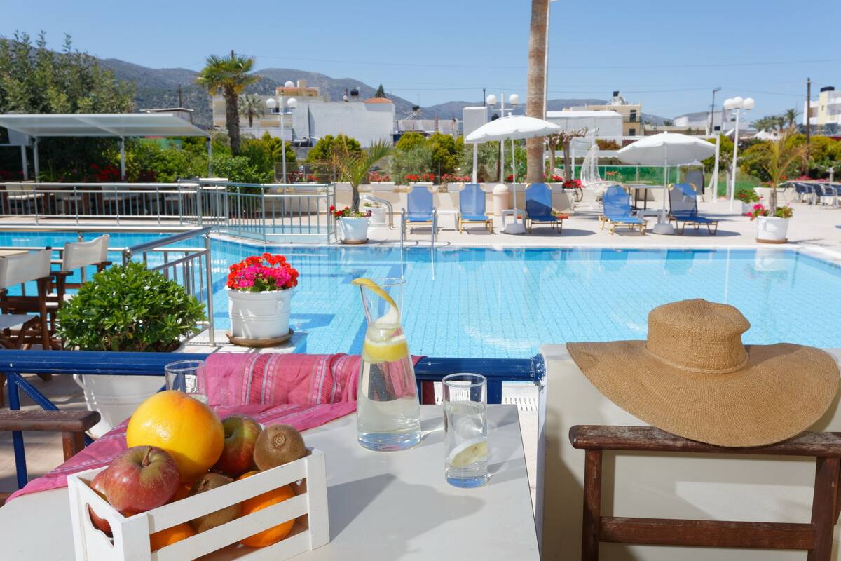 Uma área à beira da piscina com espreguiçadeiras, uma mesa com frutas e bebidas, e vista para o mar e edifícios ao fundo.