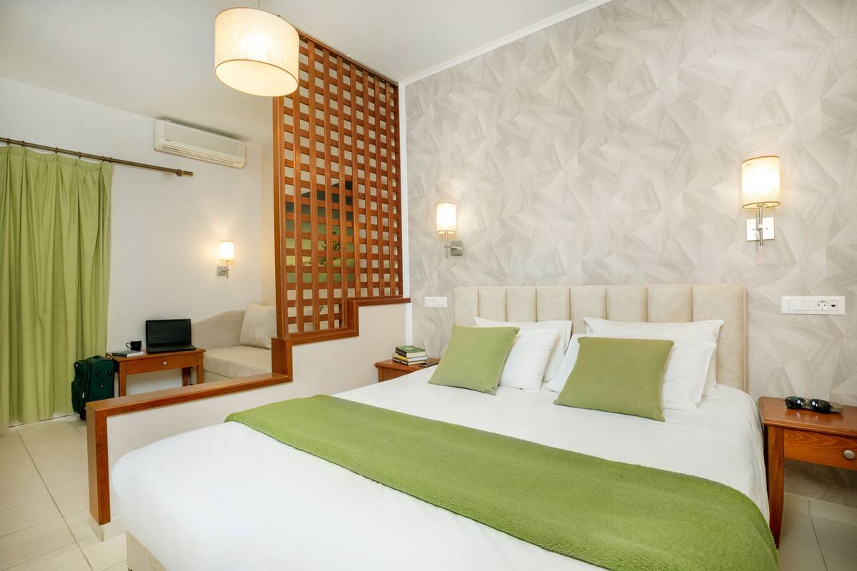 Quarto de hotel bem arrumado com uma cama, mesas de cabeceira, um laptop e uma cortina verde.