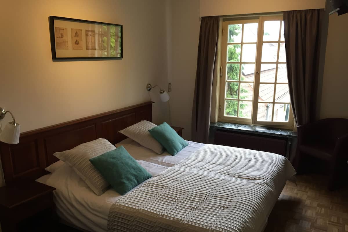 A neatly made bed with a headboard, a window with curtains, a wall-mounted lamp, and a framed picture above the bed.
