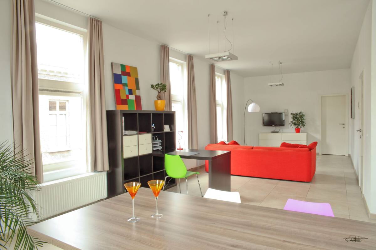 A modern living room with a wooden dining table, two cocktail glasses, a red sofa, a green chair, and a black entertainment unit.