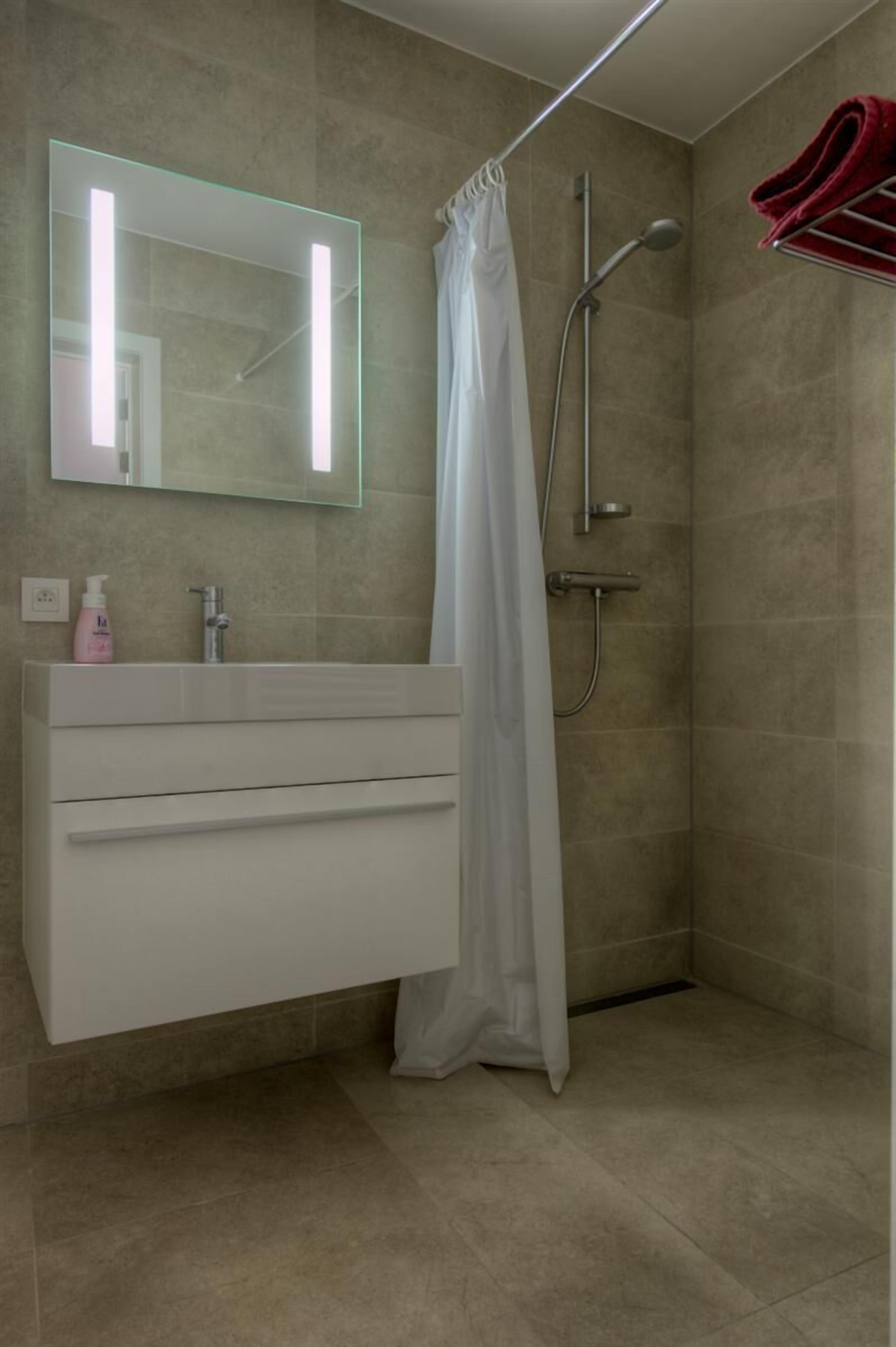 Un bagno moderno con un mobile lavabo bianco, uno specchio incassato in vetro, una doccia con tenda bianca e un portasciugamani con asciugamani rossi.