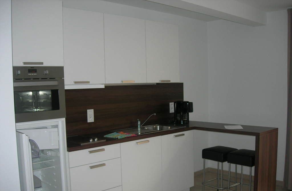 Una cucina compatta con mobili bianchi, un piano di lavoro scuro, forno integrato, macchina da caffè e due sgabelli da bar.