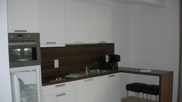Una cucina compatta con mobili bianchi, un piano di lavoro scuro, forno integrato, macchina da caffè e due sgabelli da bar.
