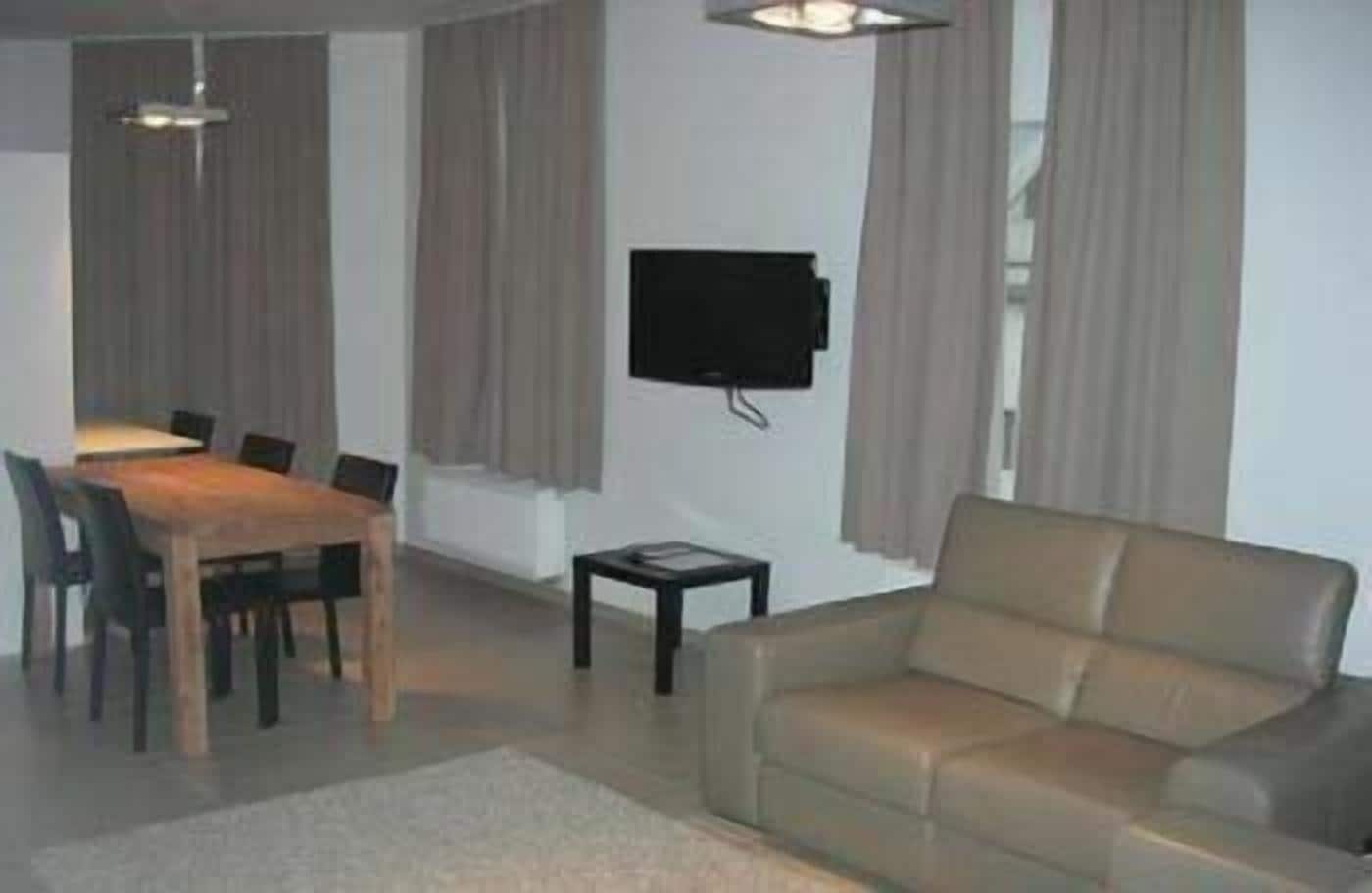 A living room with a sofa, a dining table with chairs, a TV mounted on the wall, and a small table.