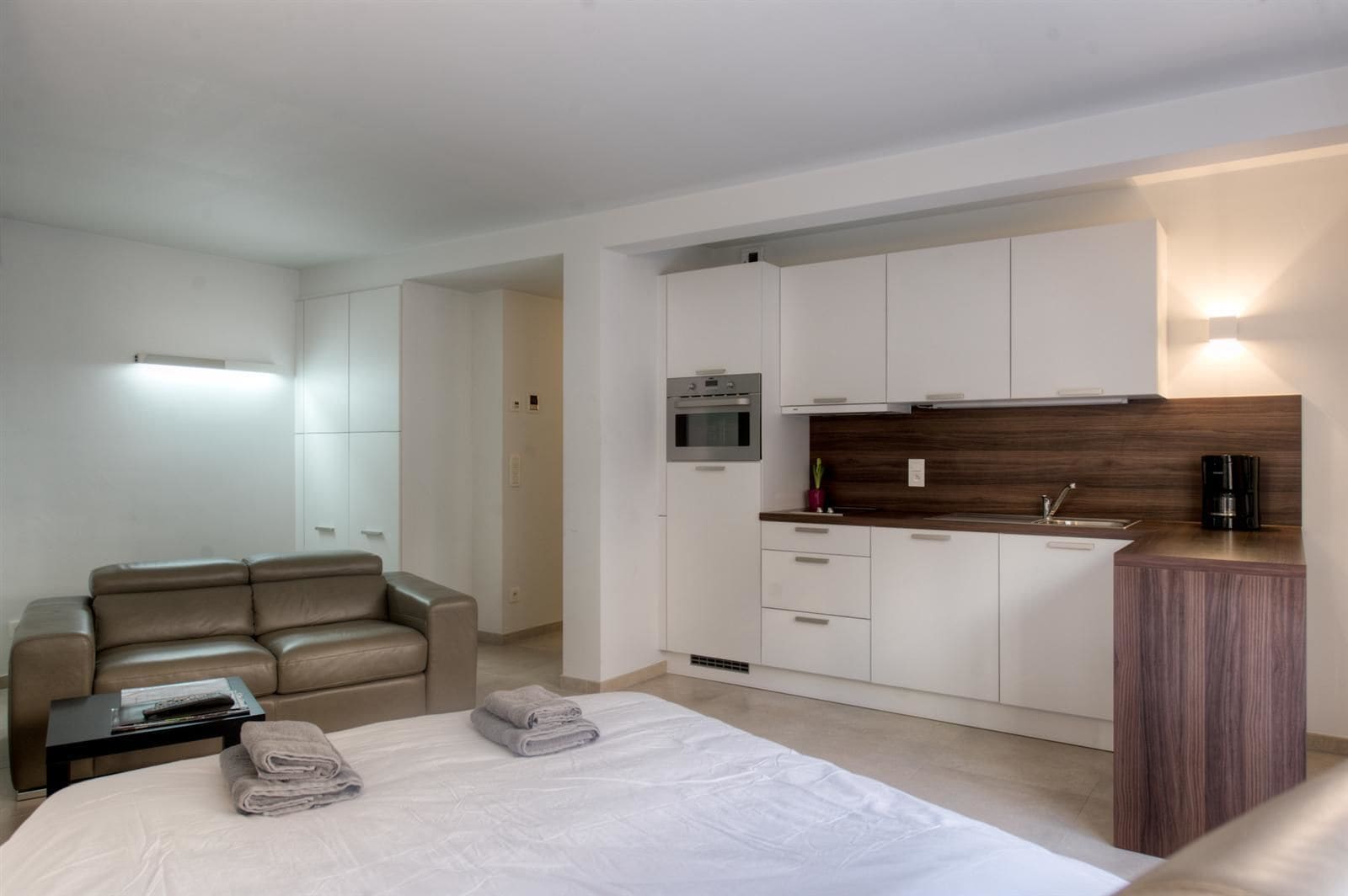 Una habitación de hotel moderna con una cama, un sofá, una pequeña mesa con una revista y una zona de cocina con microondas, horno y cafetera.