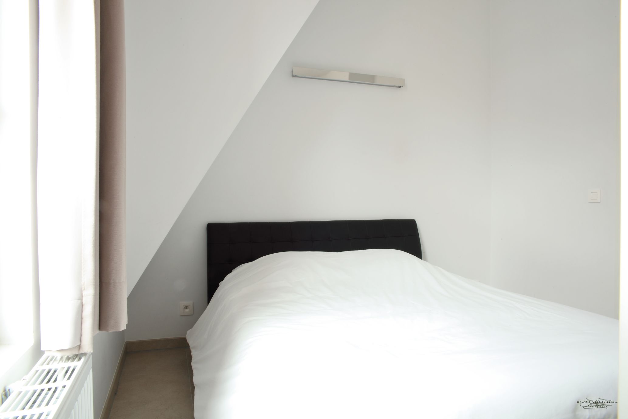 A small, neatly made bed in a corner room with a sloped ceiling and a window with curtains.