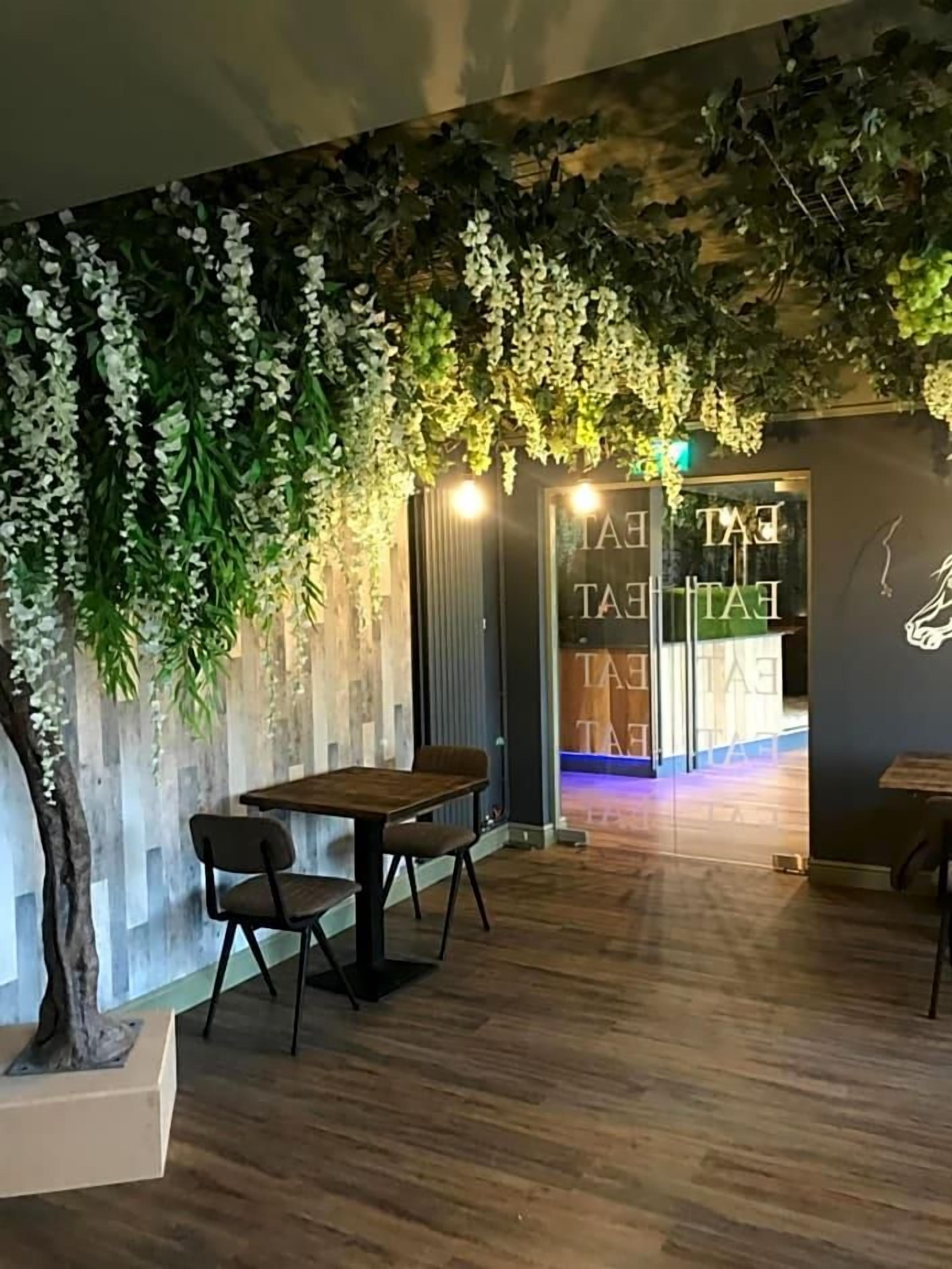 A room with a wooden floor, a table with chairs, a wall with hanging greenery, and a door with a sign.