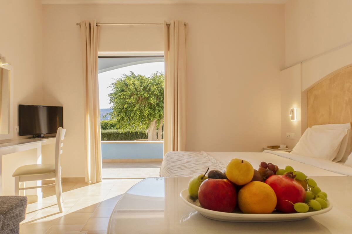 Une chambre d’hôtel avec un lit, une table à manger agrémentée d’un panier de fruits, une télévision et une vue sur la verdure environnante.