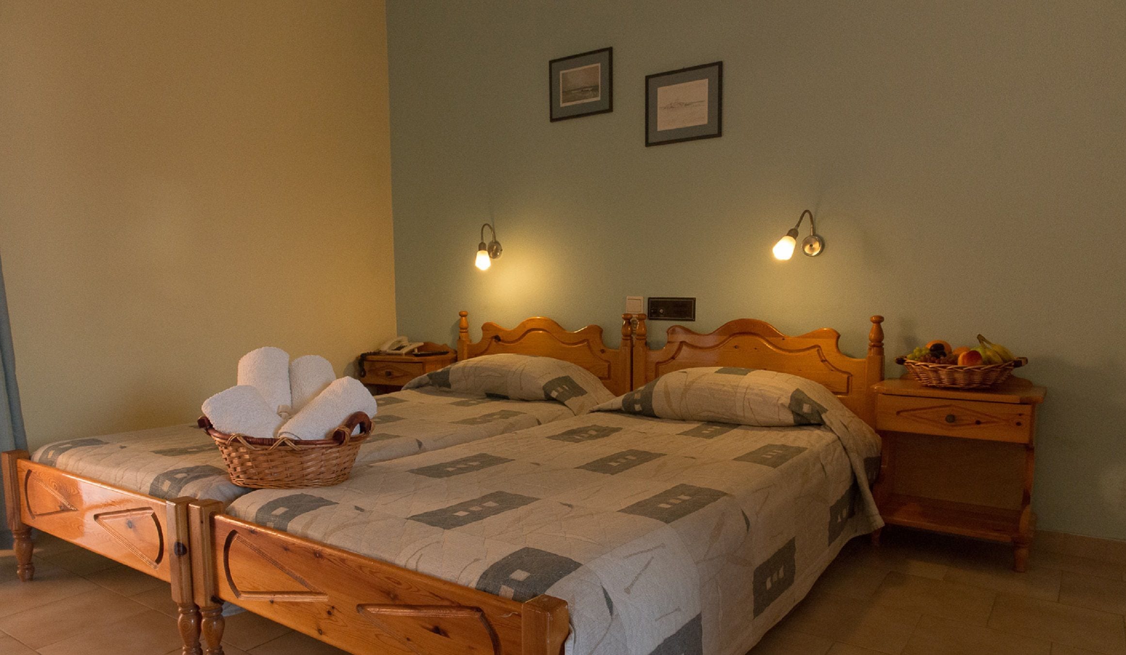 A double bed with a wooden headboard, two bedside tables, a wall-mounted light fixture, and a basket of towels.