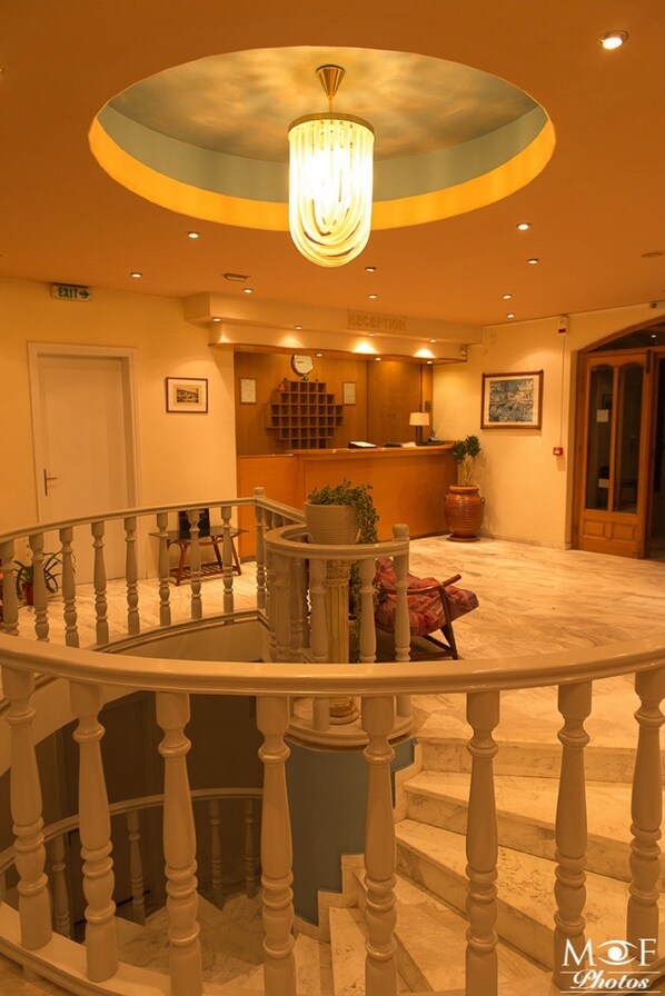 A hotel lobby with a reception desk, a chandelier, a staircase with a railing, and a potted plant.