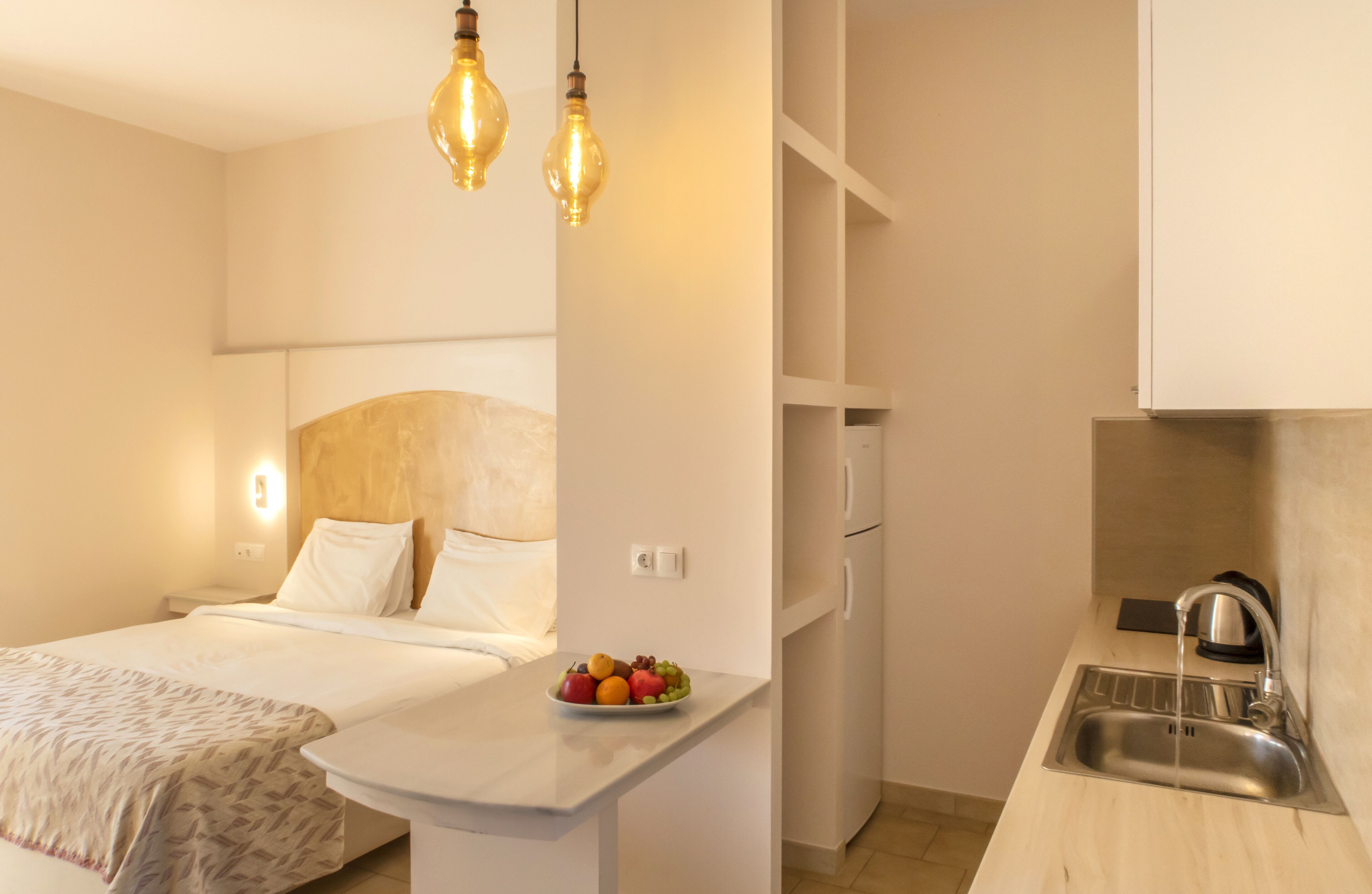 Una camera d'albergo compatta con un letto, comodino, vassoio di frutta e angolo cottura con lavello e fornelli.
