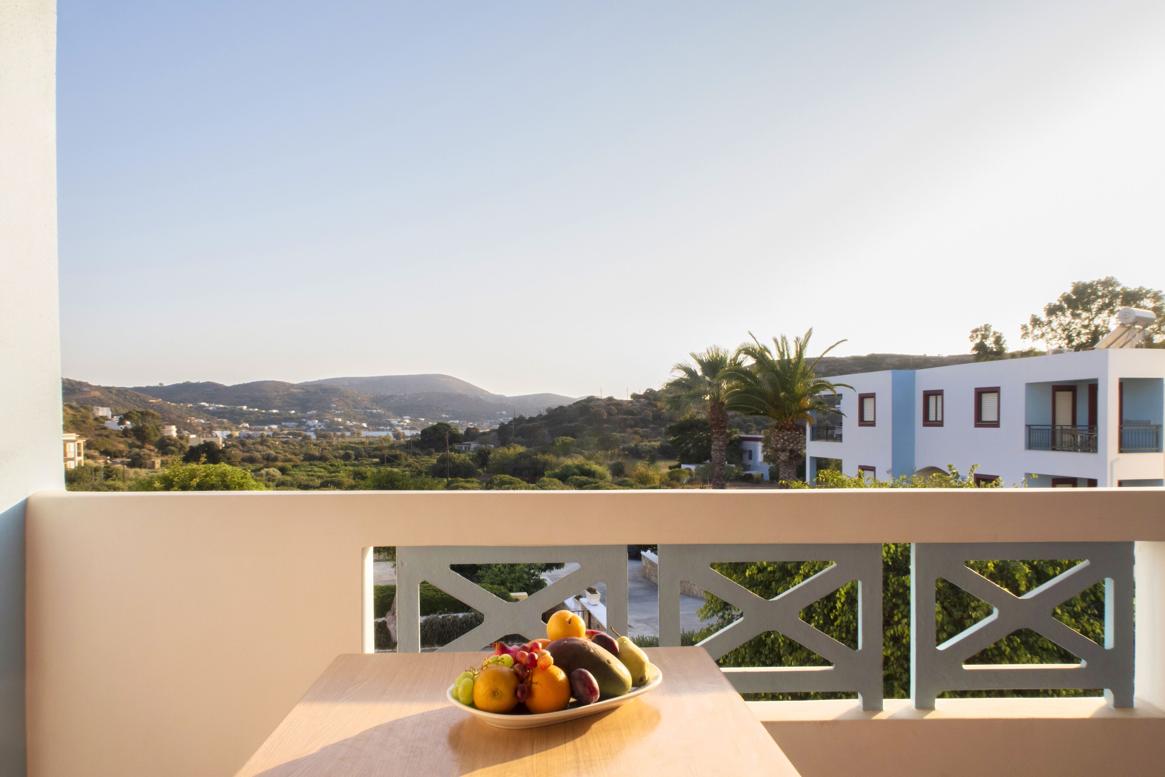 Varanda com uma mesa que suporta uma tigela de frutas, com vista para a paisagem costeira com edifícios e colinas.