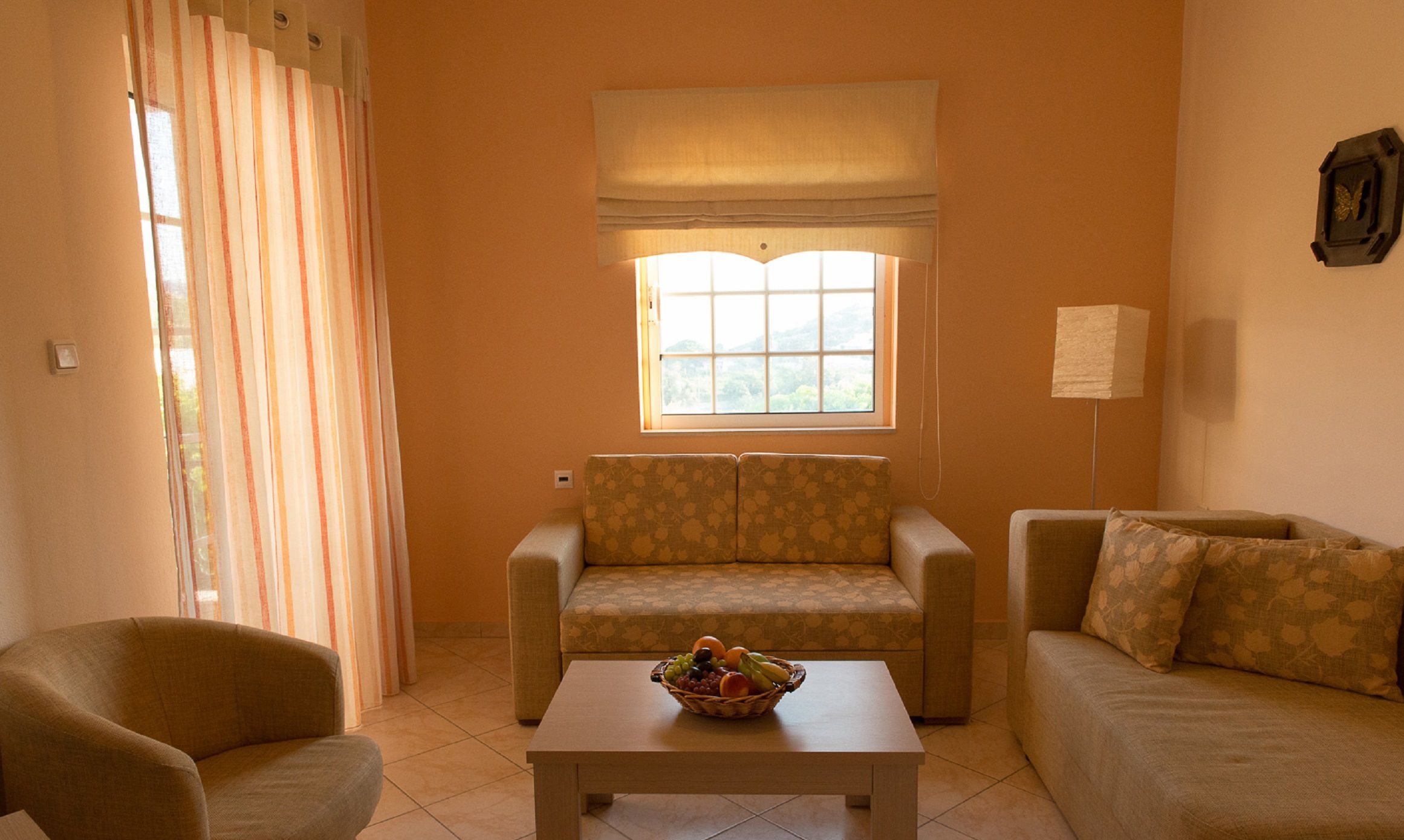 A living room with a sofa, a chair, a table with a fruit basket, a window with a view, and a lamp.