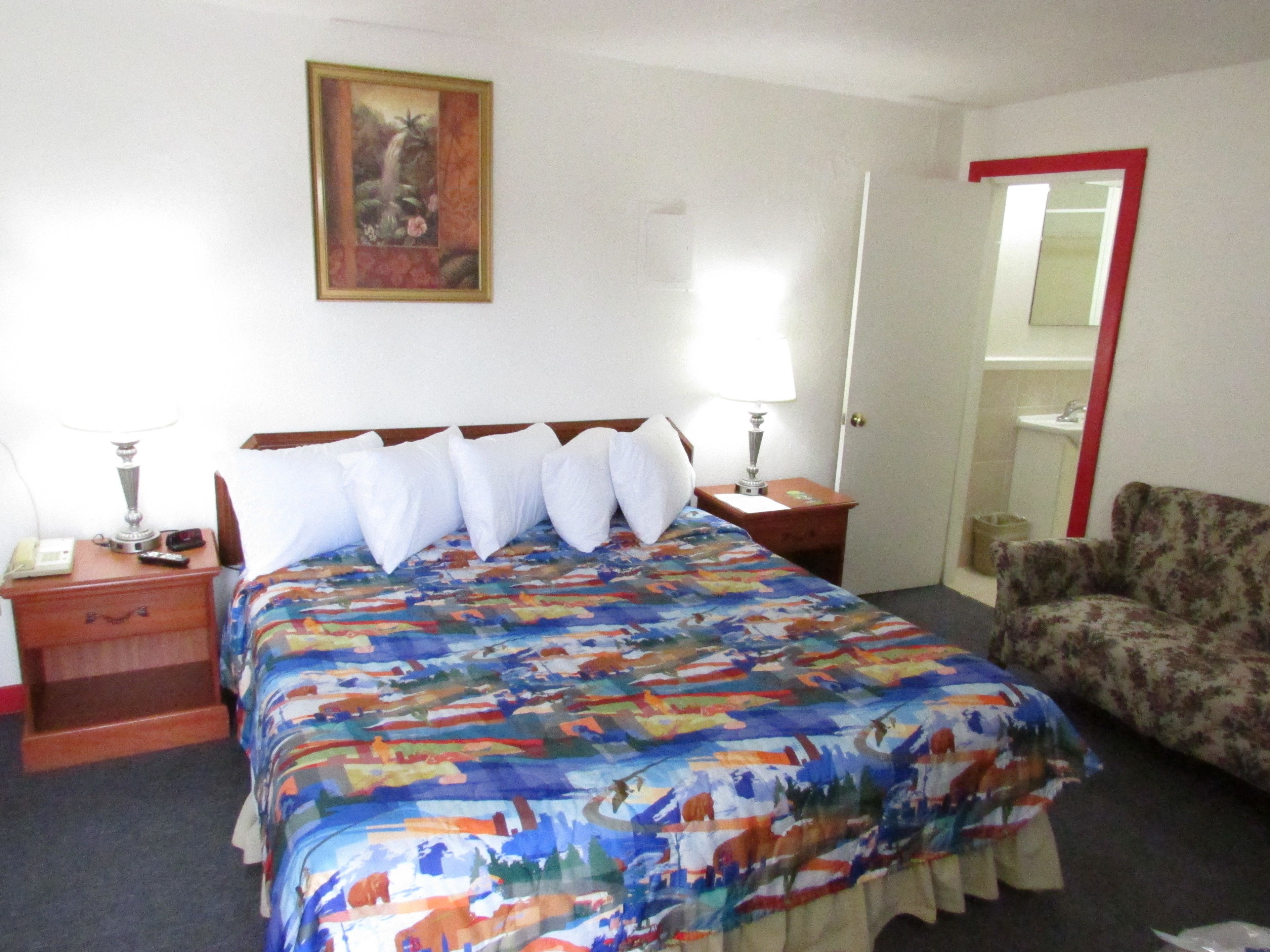 Une chambre d’hôtel avec un lit, deux tables de chevet, une chaise, un tableau au mur et une porte avec un cadre rouge.