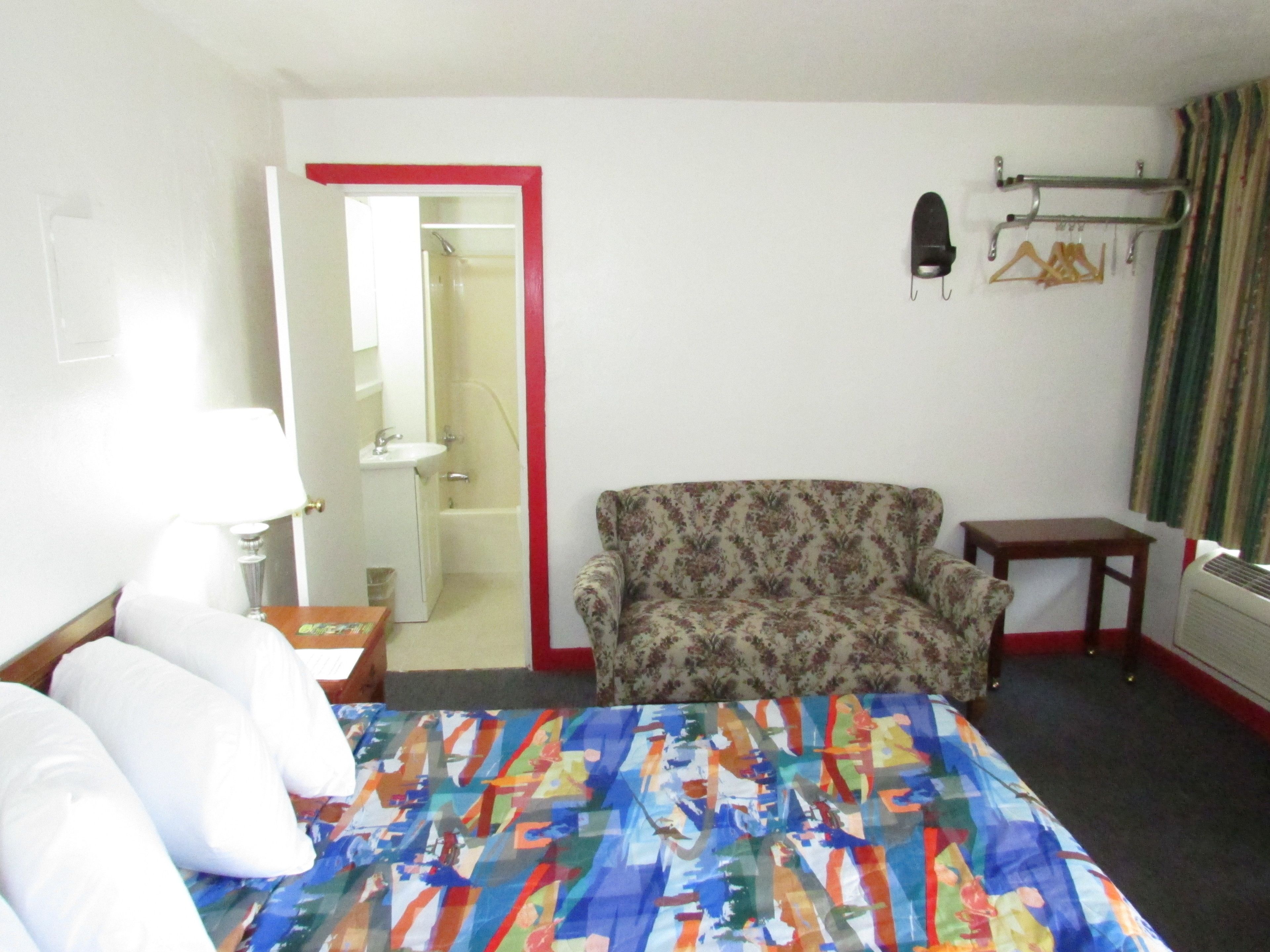 Une chambre d’hôtel comprenant un lit, une chaise, une petite table, une lampe, un miroir et un espace douche.