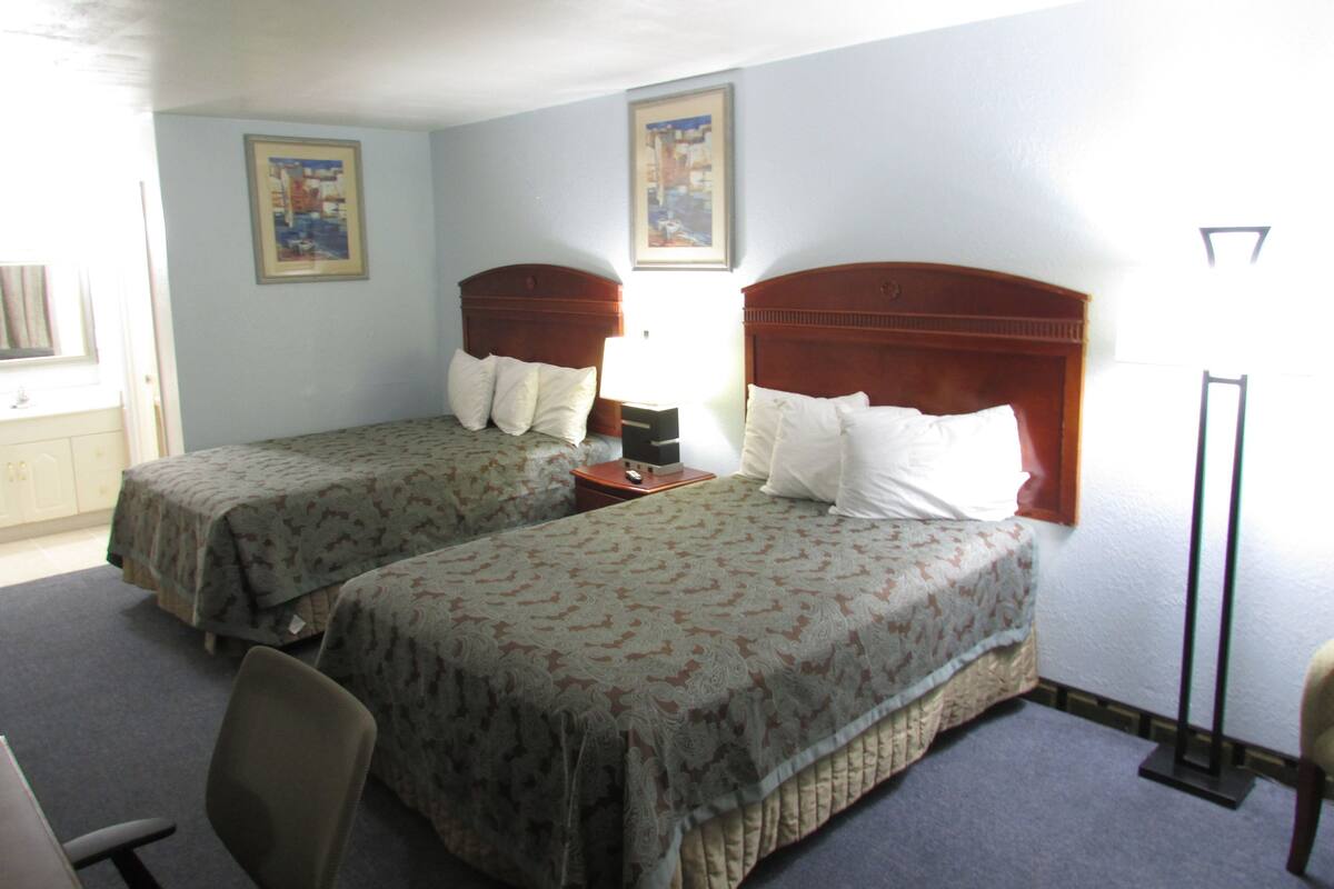 A hotel room with two single beds, a desk, a chair, and a nightstand with a lamp.