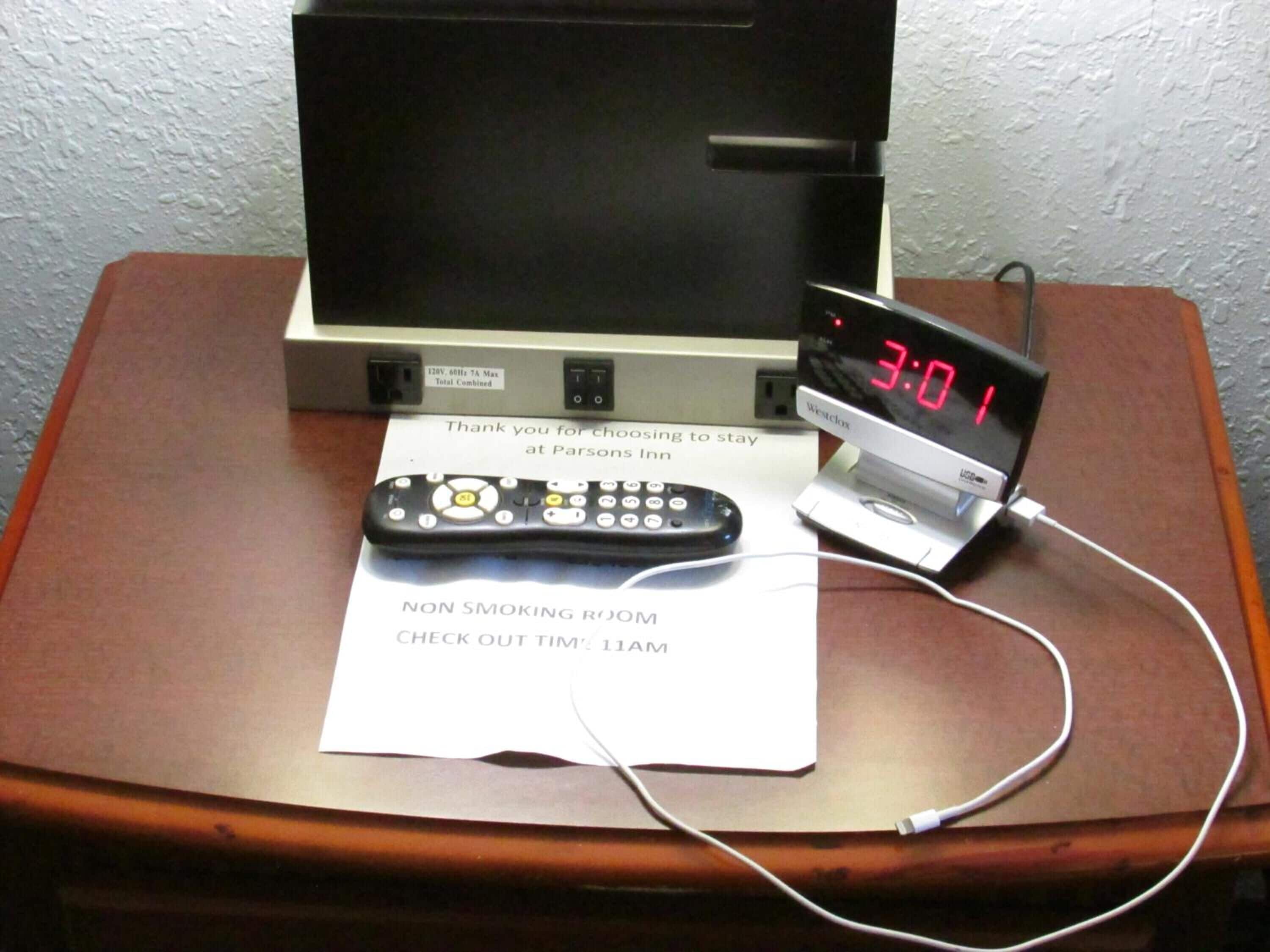 A hotel room with a digital clock displaying 3:01, a remote control, a non-smoking room sign, and a check-out time notice.