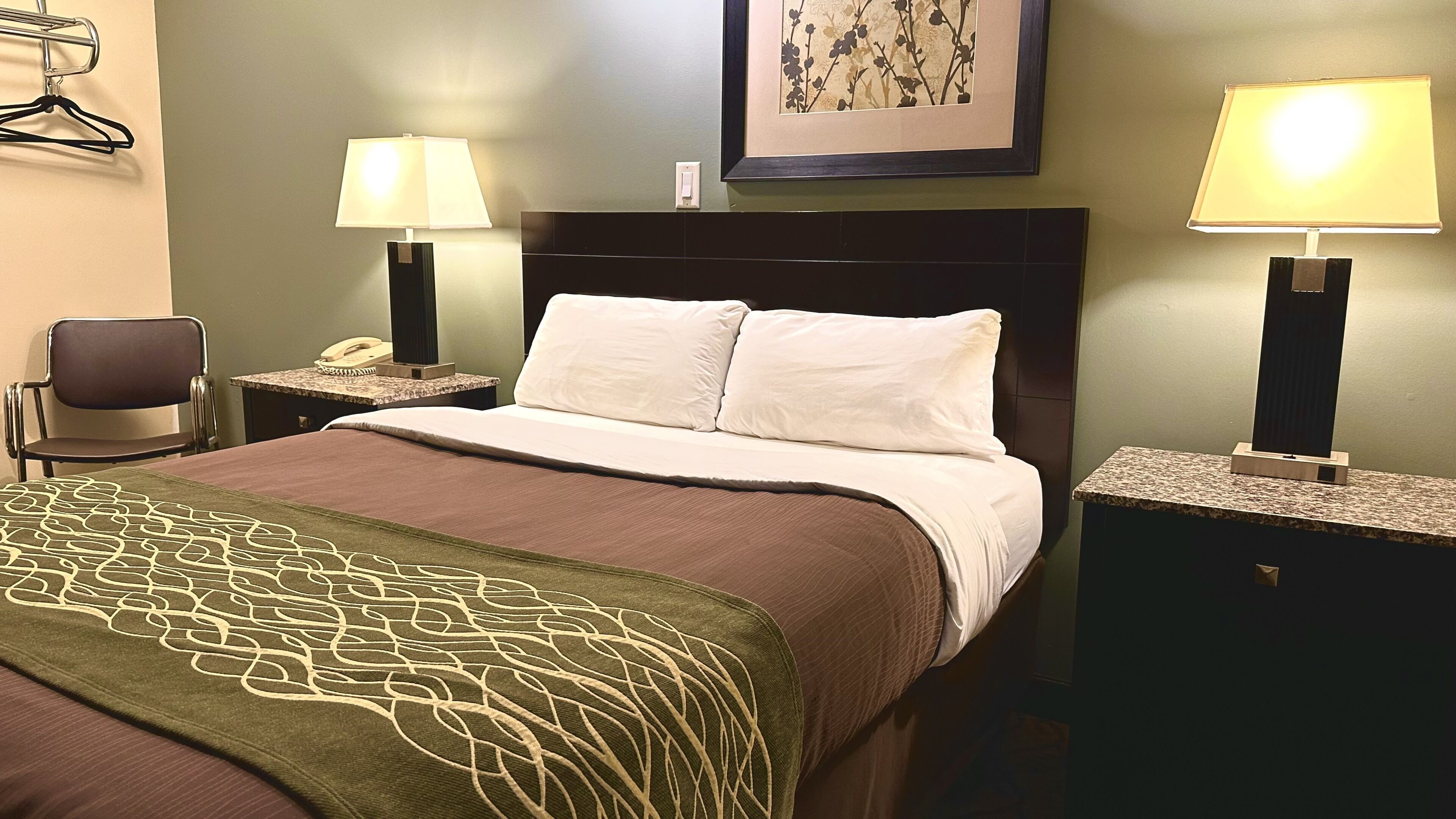 A hotel room with a bed, two bedside tables with lamps, a chair, a telephone, and a framed picture on the wall.