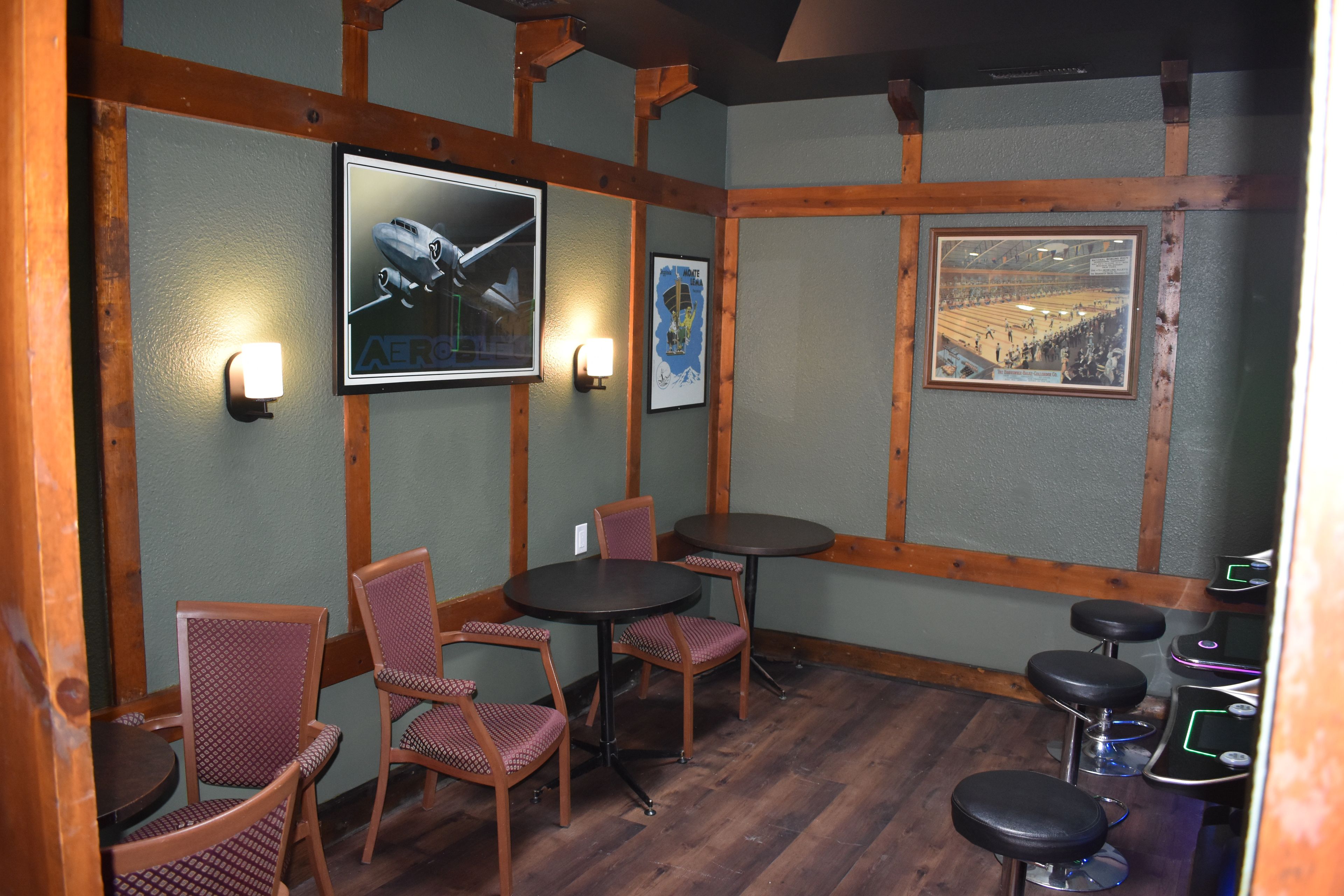 Een kamer met houten balken, een houten vloer en een wand met ingelijste foto's. Er staan stoelen en tafels.
