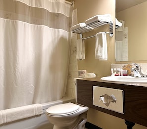 A bathroom with a shower curtain, a towel rack with towels, a toilet, a sink with toiletries, and a mirror. - Lacombe Motor Inn (Lacombe)