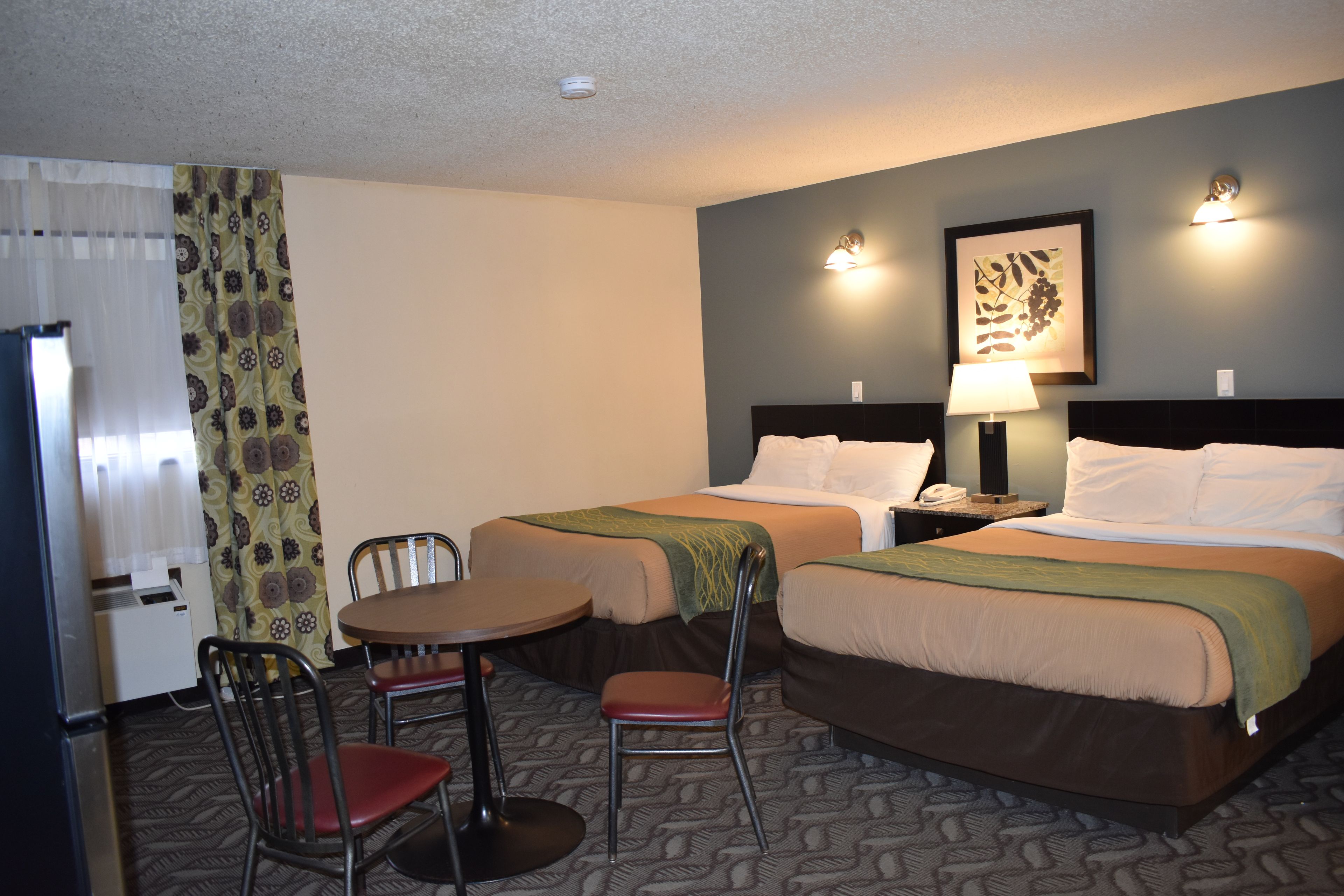 A hotel room with two beds, a small table with two chairs, a desk with a telephone, and a wall-mounted lamp.