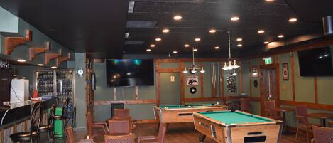 A bar area with pool tables, chairs, and a bar counter with a clock and bottles.
