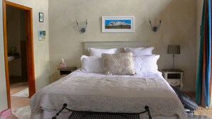Una cama bien tendida con sábanas blancas, un cuadro enmarcado de una montaña en la pared y una lámpara en una mesita de noche.