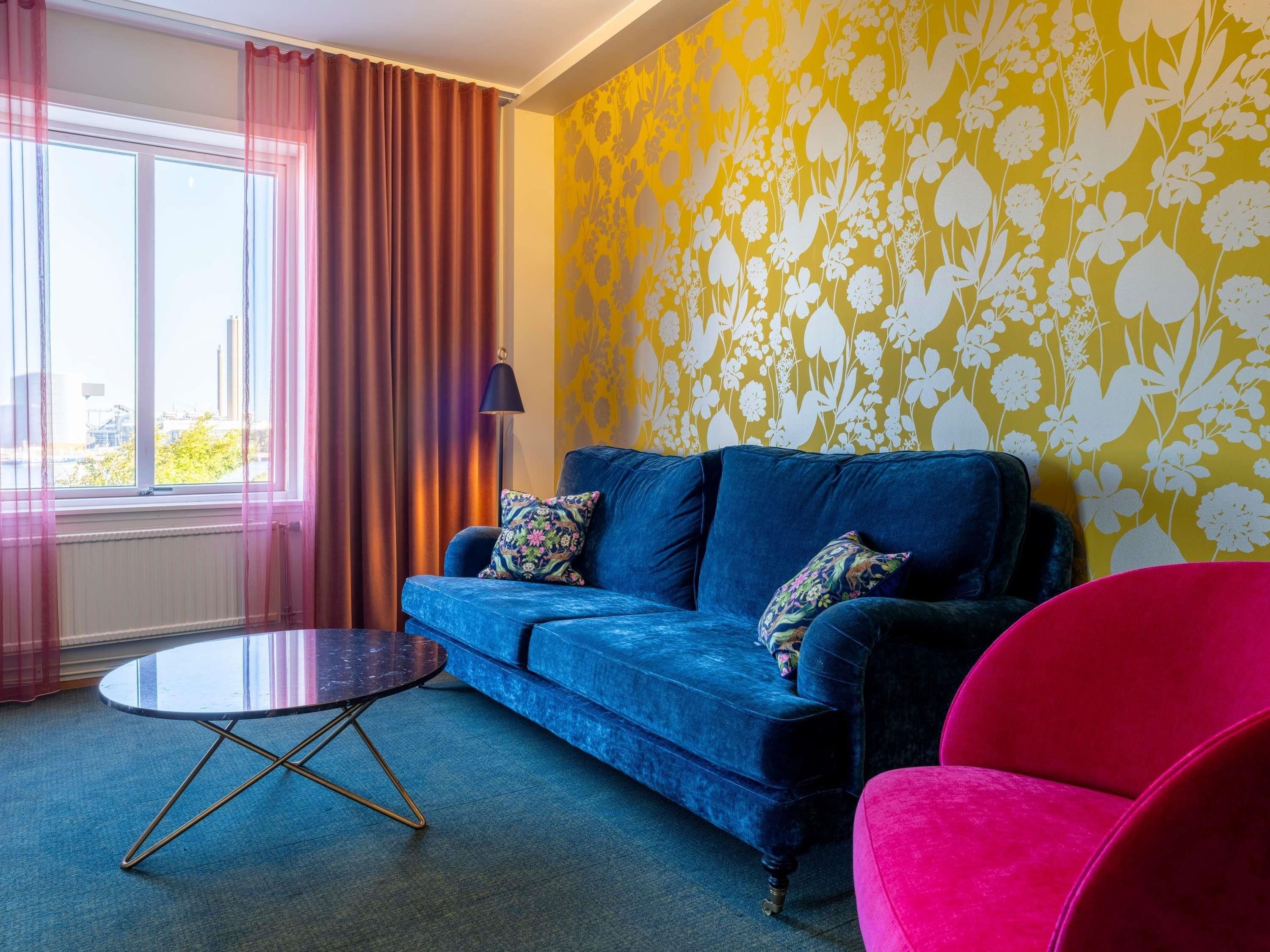 A room with a blue sofa, a round table, a red chair, and a yellow floral wallpaper.