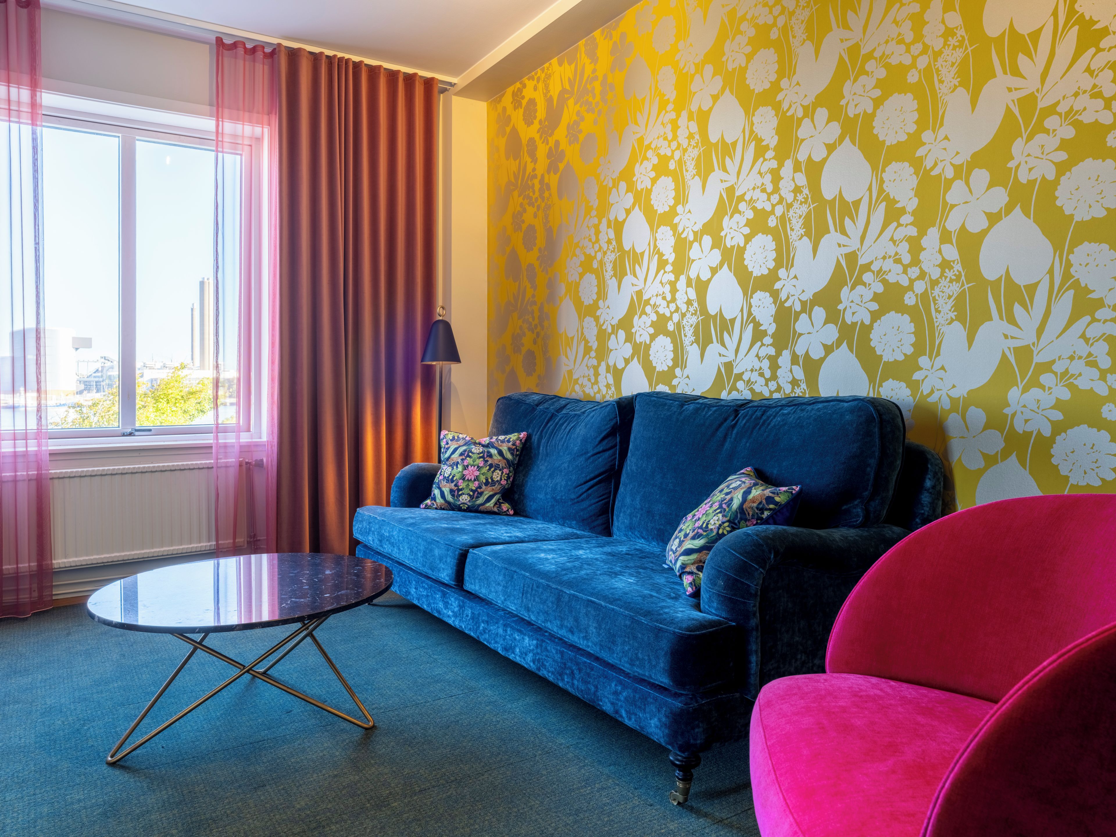 A room with a blue sofa, a round coffee table, a red chair, and a window with curtains.