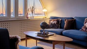 A modern living room with a blue sofa, a wooden coffee table, and a view of the harbor with cranes.