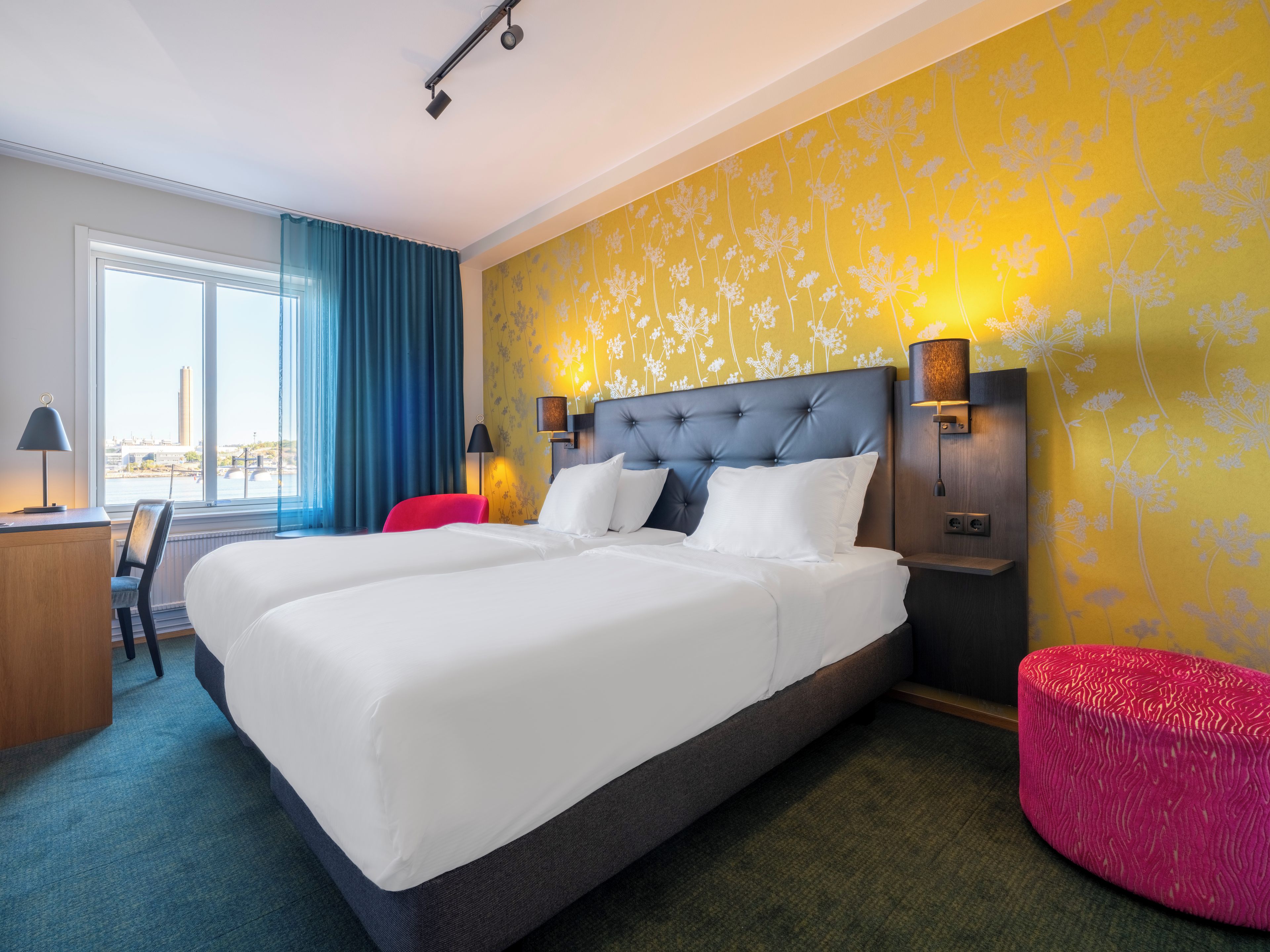 A hotel room with a large bed, two bedside lamps, a desk with a chair, and a view of the city.