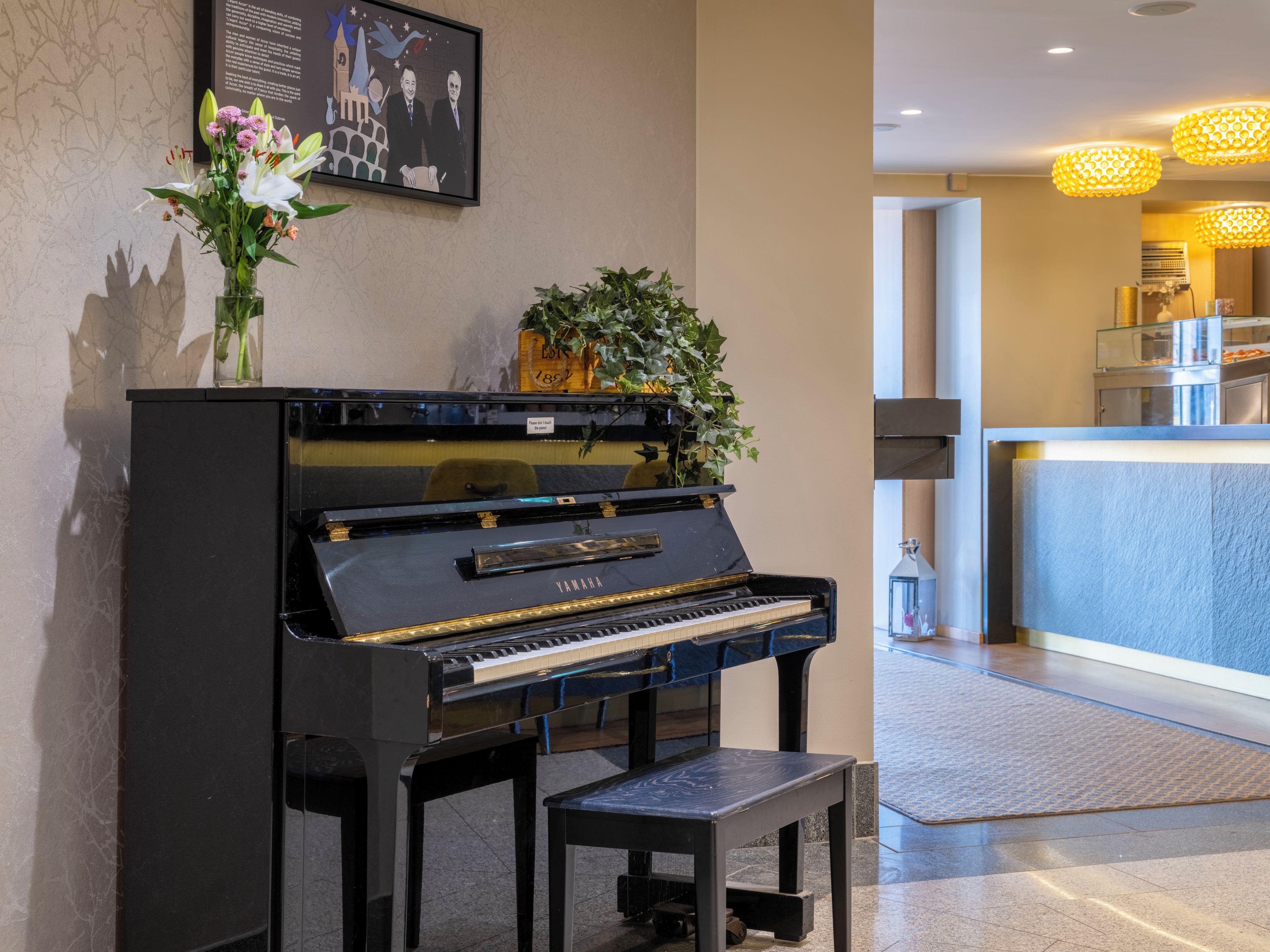 A Yamaha grand piano in a lobby area with a framed picture above it, a potted plant, and a small table in front.