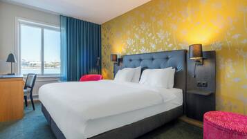 A hotel room with a large bed, a desk, a chair, a red ottoman, and a view of the city through the window.
