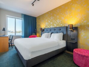 A hotel room with a large bed, a desk, a chair, a red ottoman, and a view of the city through the window. - Mercure Gothenburg (Vastra Frolunda)