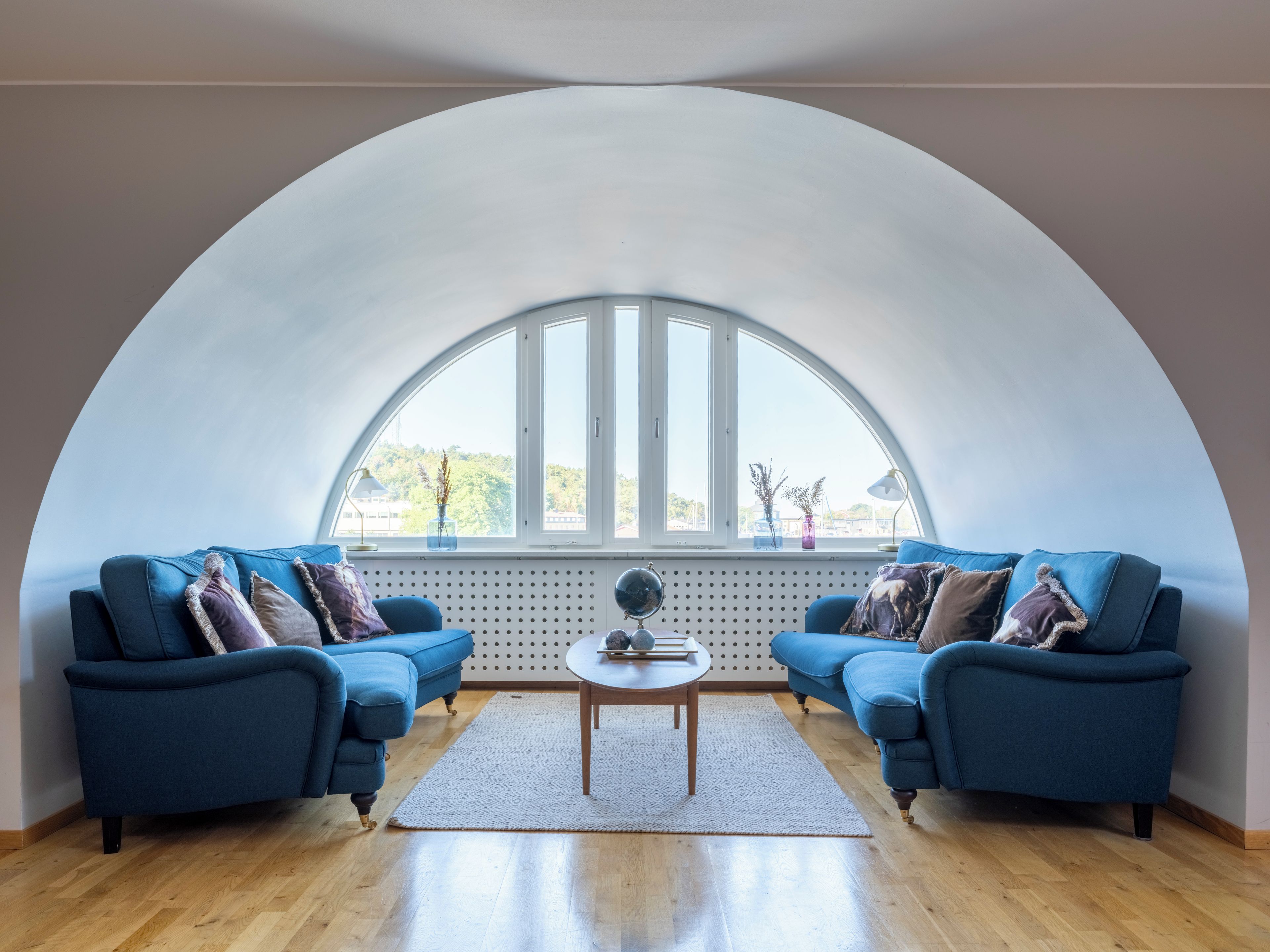 Una habitación con techo semicircular, dos sofás azules, una mesa redonda de madera y un ventanal con vistas a la vegetación.