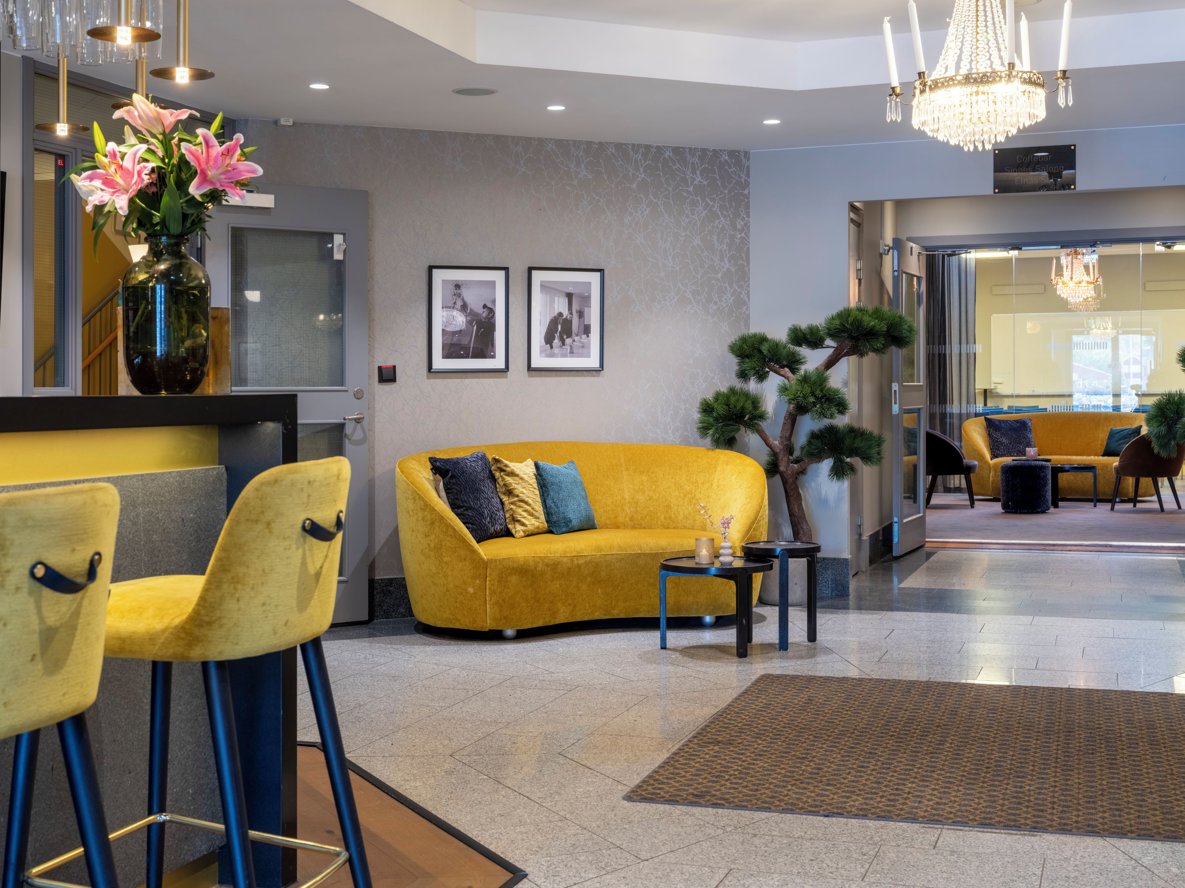 A modern lobby with a yellow sofa, two bar stools, a small table, and a decorative tree.