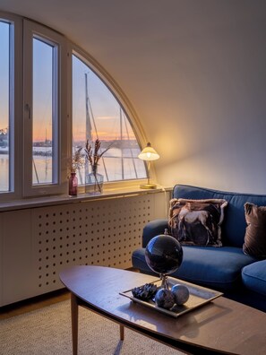 A cozy living room with a curved window overlooking a bridge, a blue sofa with decorative pillows, a coffee table with a globe and decorative items, and a lamp providing warm light. - Mercure Gothenburg (Vastra Frolunda)