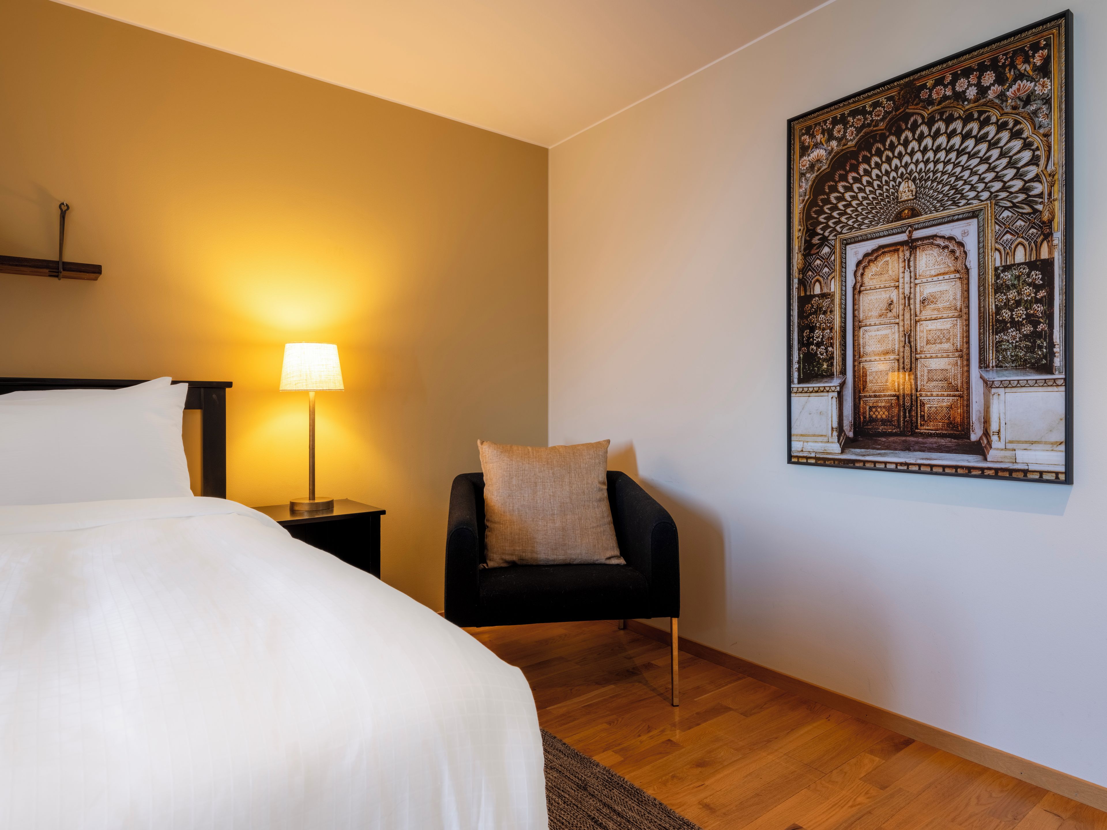 A hotel room with a bed, a nightstand with a lamp, a chair, and a wall-mounted artwork.