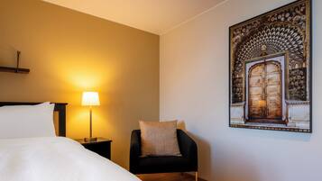 A hotel room with a bed, a nightstand with a lamp, a chair, and a wall-mounted artwork.