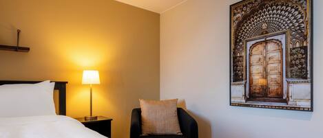 Habitación de hotel con cama, mesita de noche con lámpara, una silla y una obra de arte colgada en la pared.