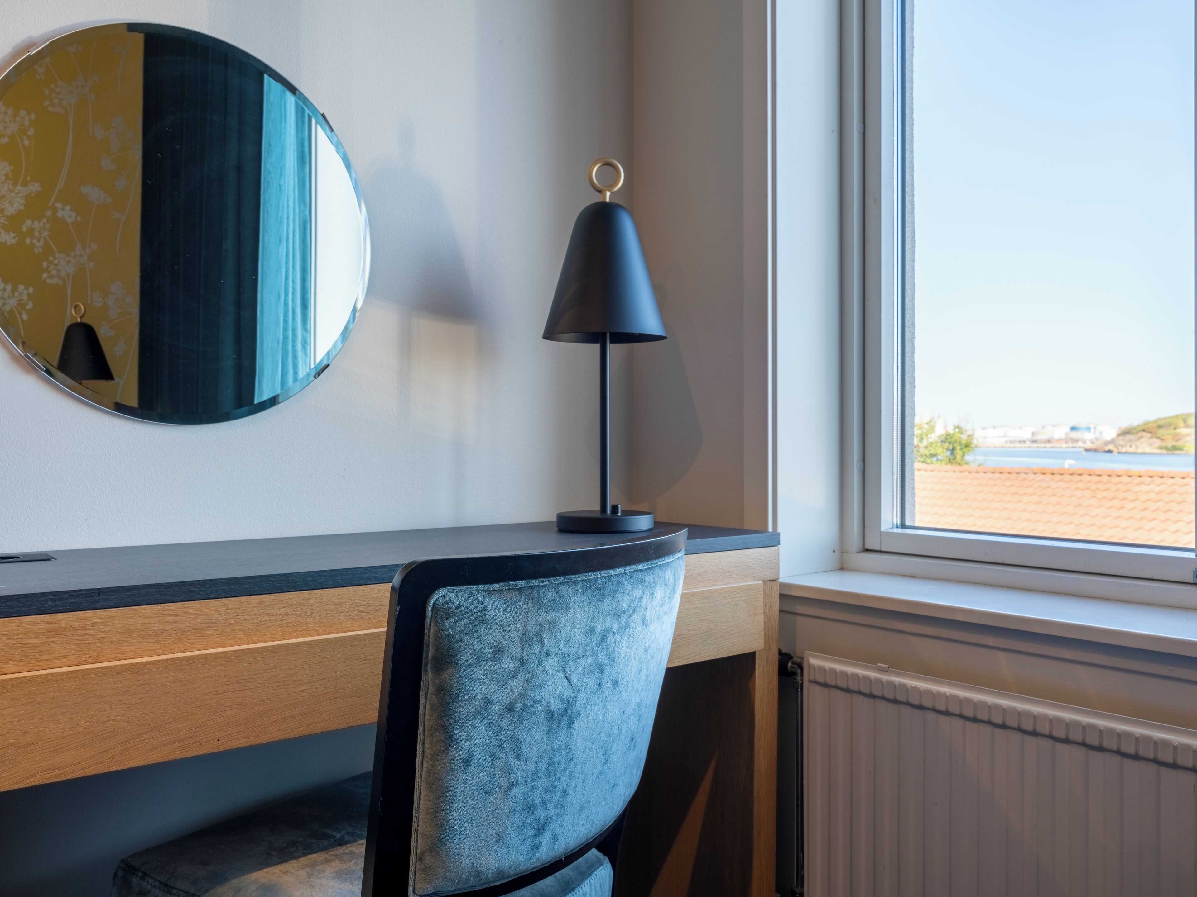 A room with a desk, a chair, a lamp, a round mirror, and a window with a view.