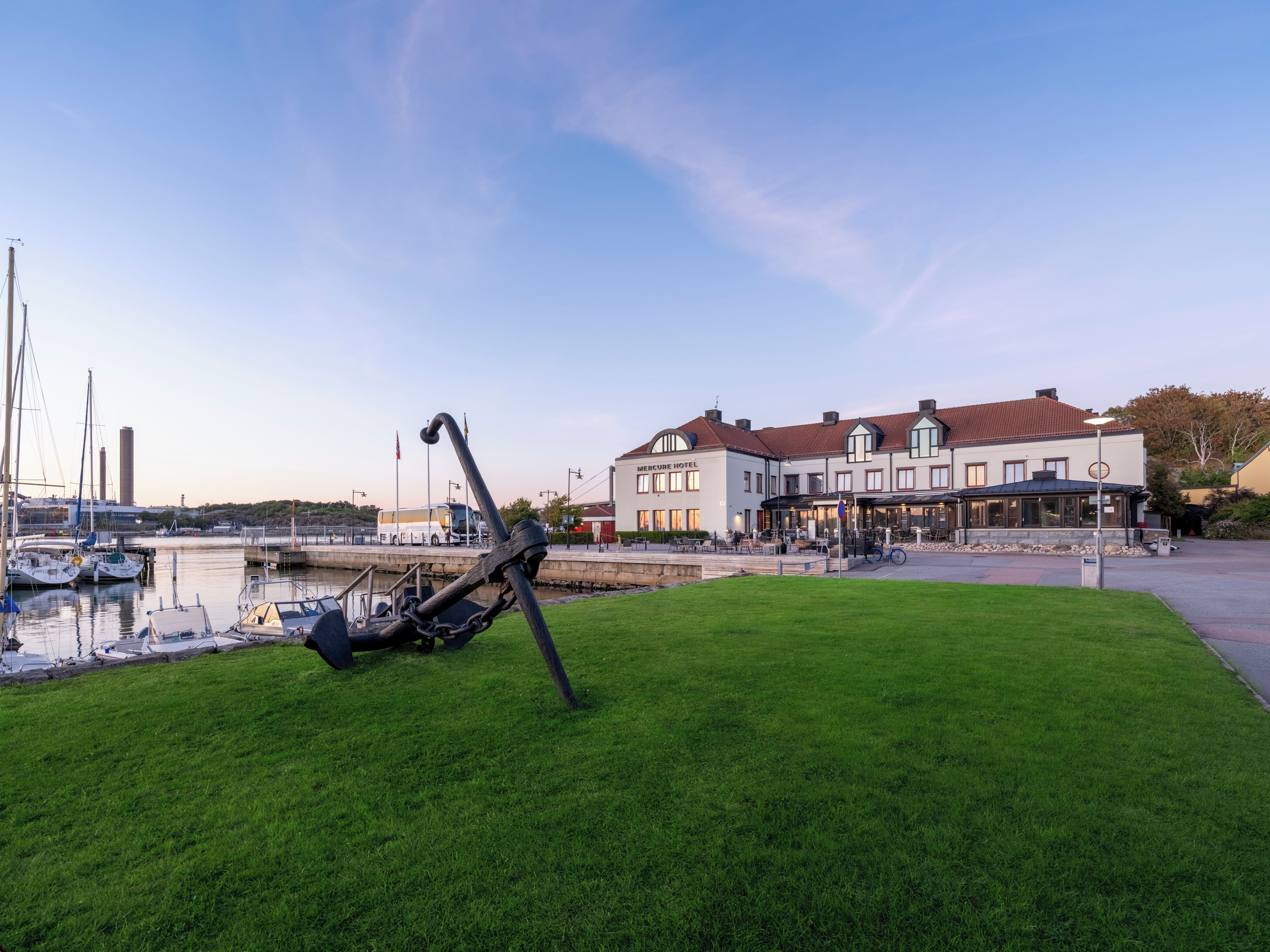 A grassy area with a large metal sculpture in the foreground, a marina with boats, and a hotel with a red roof in the background.