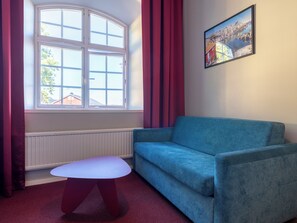 A room with a blue sofa, a small white table, a window with curtains, and a framed picture on the wall. - Mercure Gothenburg (Vastra Frolunda)