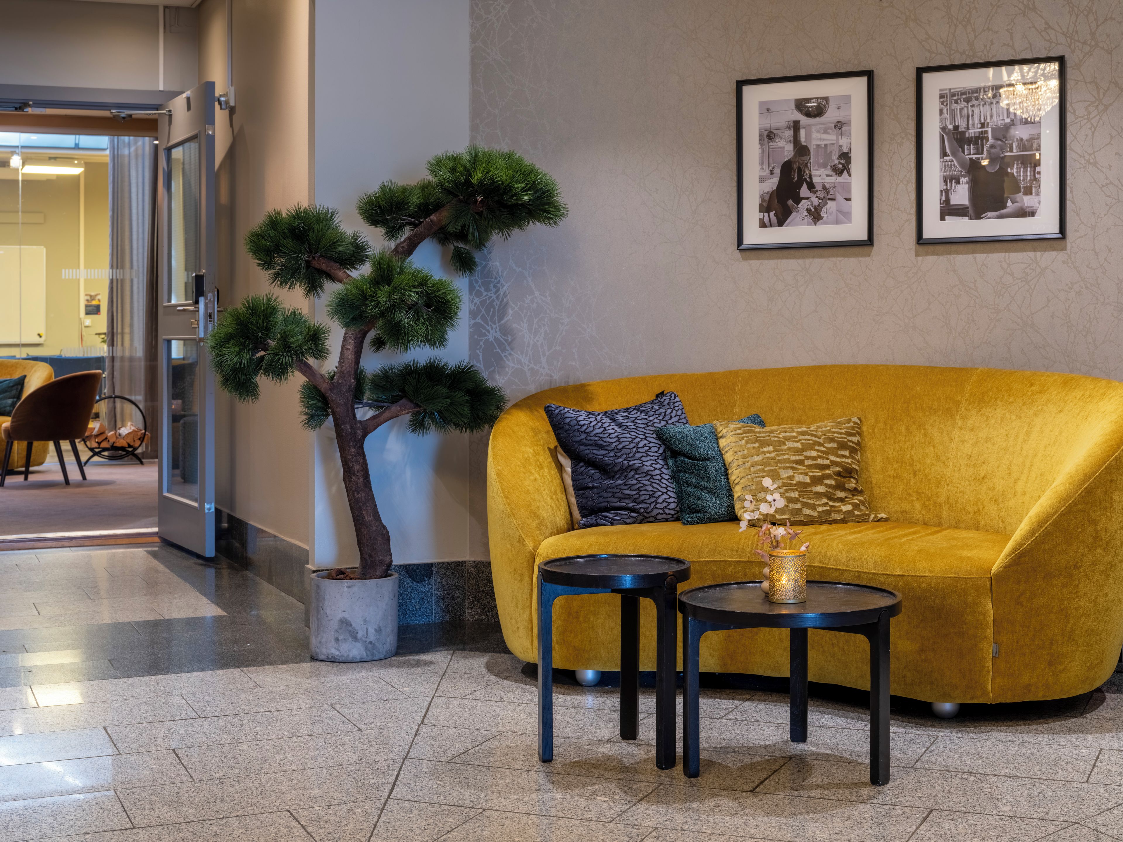Un intérieur moderne avec un canapé jaune, deux petites tables, un arbre en pot et deux photographies encadrées accrochées au mur.