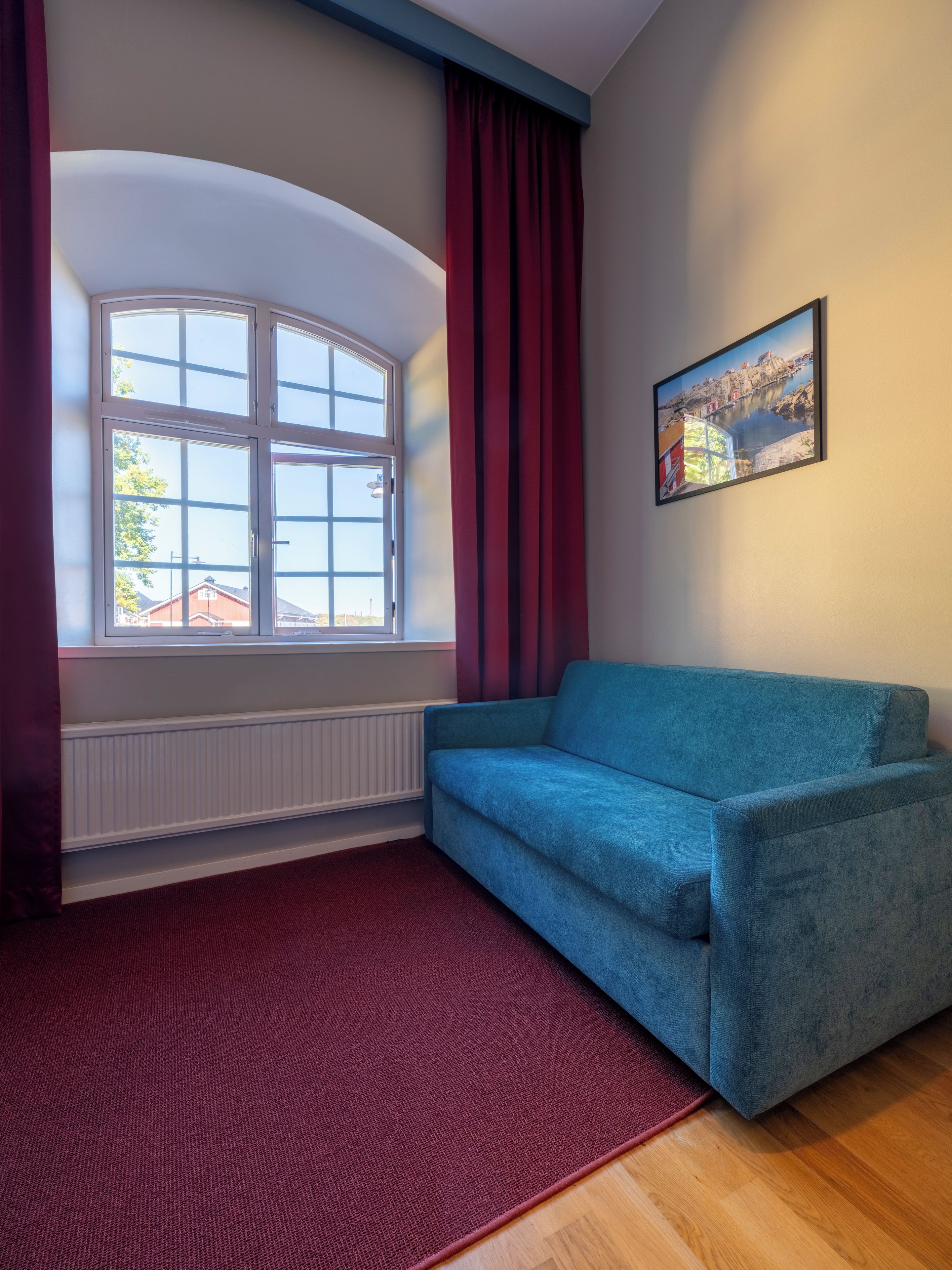 A room with a blue sofa, a red curtain, a window with a view of buildings, and a framed picture on the wall.