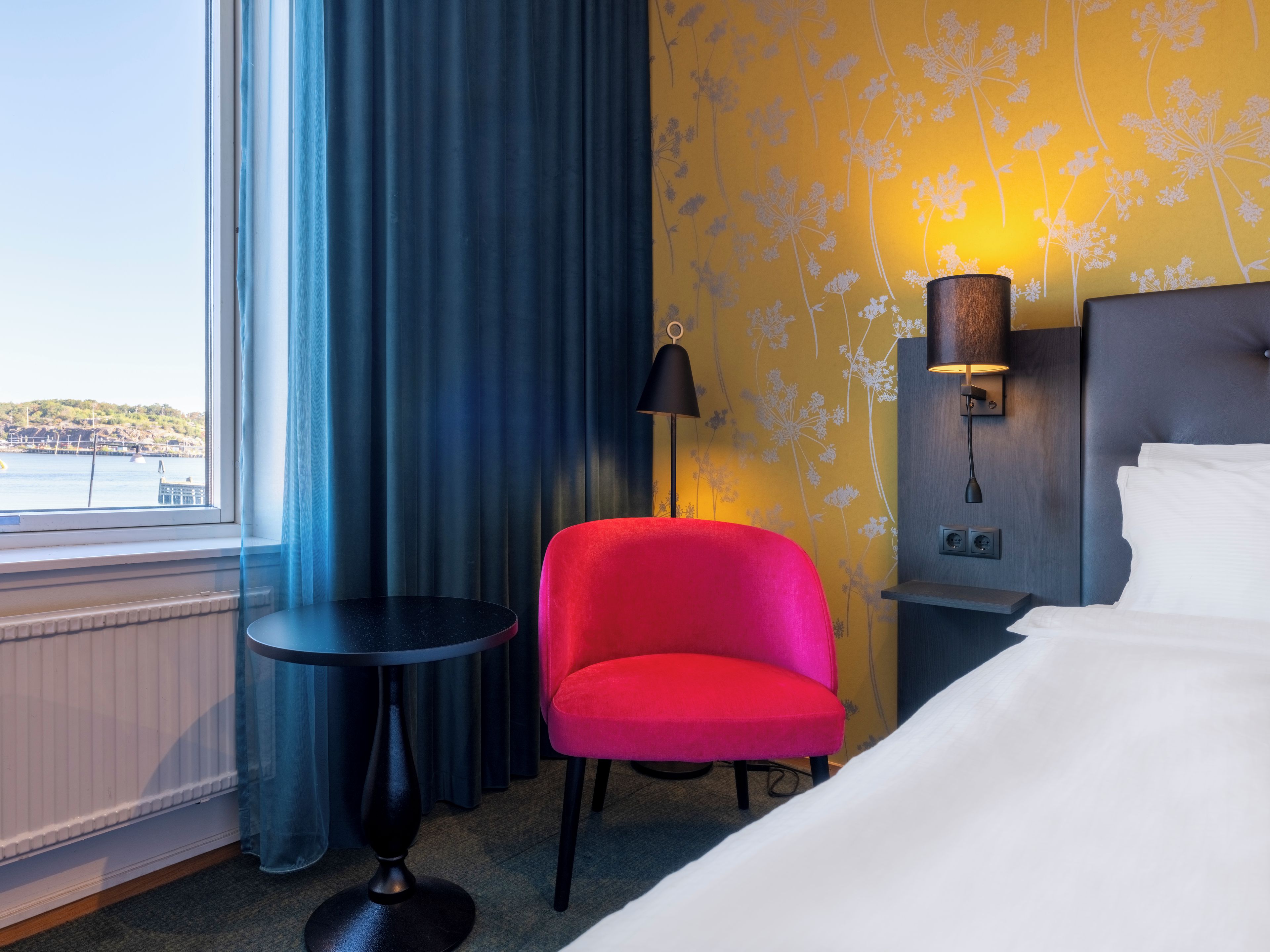 A hotel room with a bed, a red chair, a small table, a lamp, and a view of the sea through a window.