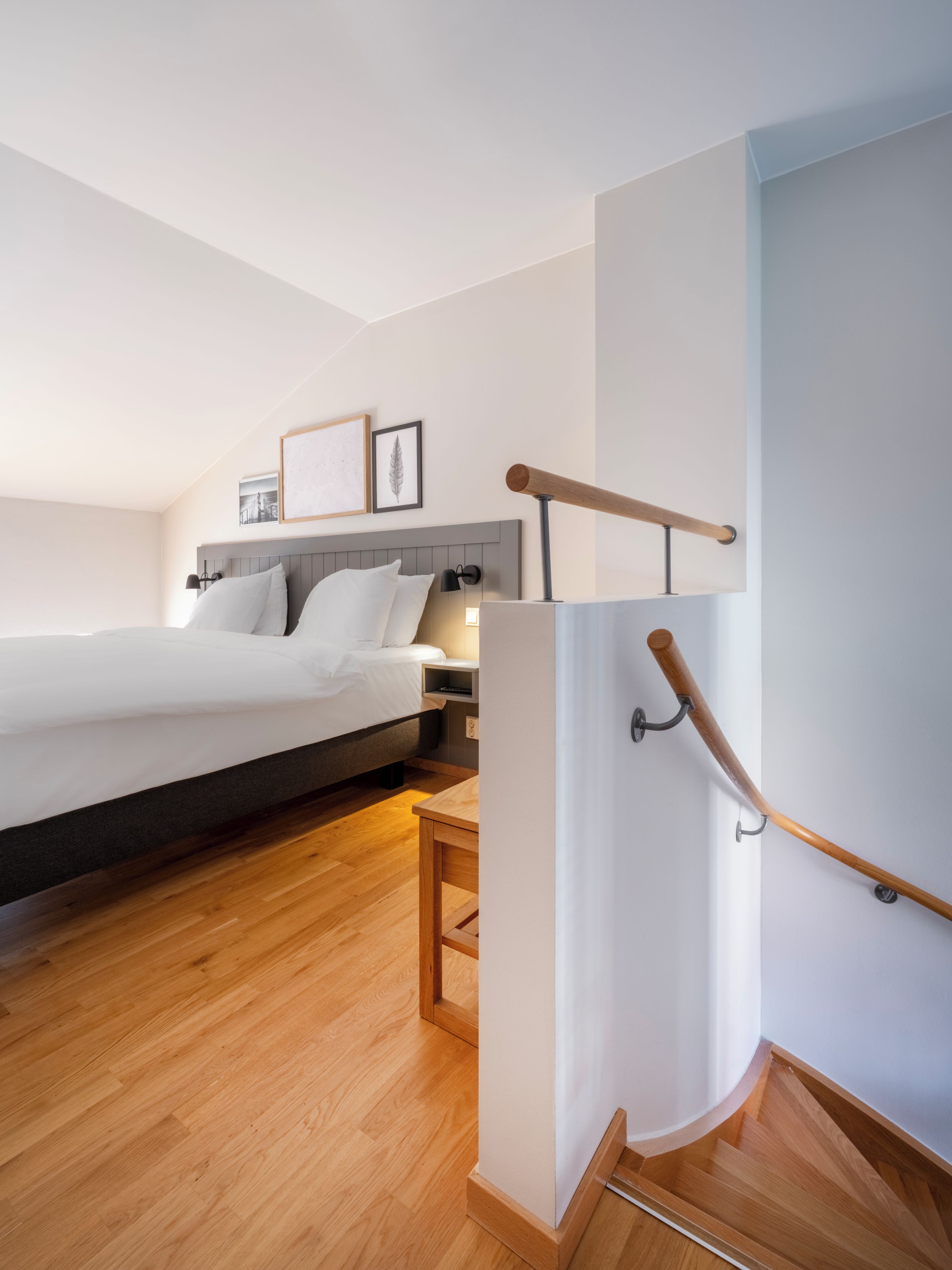 A modern hotel room with a wooden floor, a bed with white linens, a nightstand with a lamp, and a framed picture on the wall.